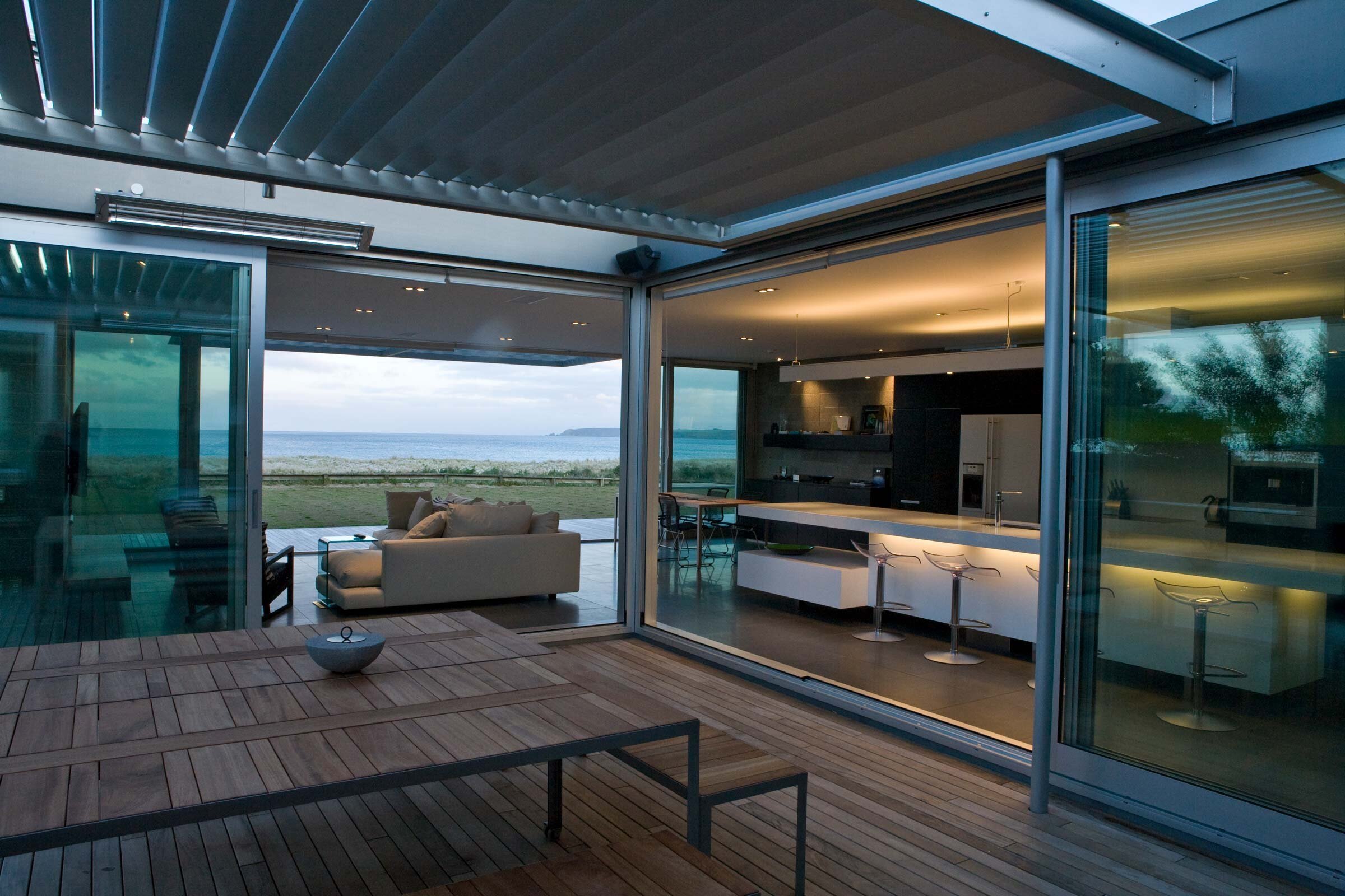 Modern open-concept home with a kitchen and living area, featuring large glass walls and ocean views at dusk. Stylish outdoor dining space.