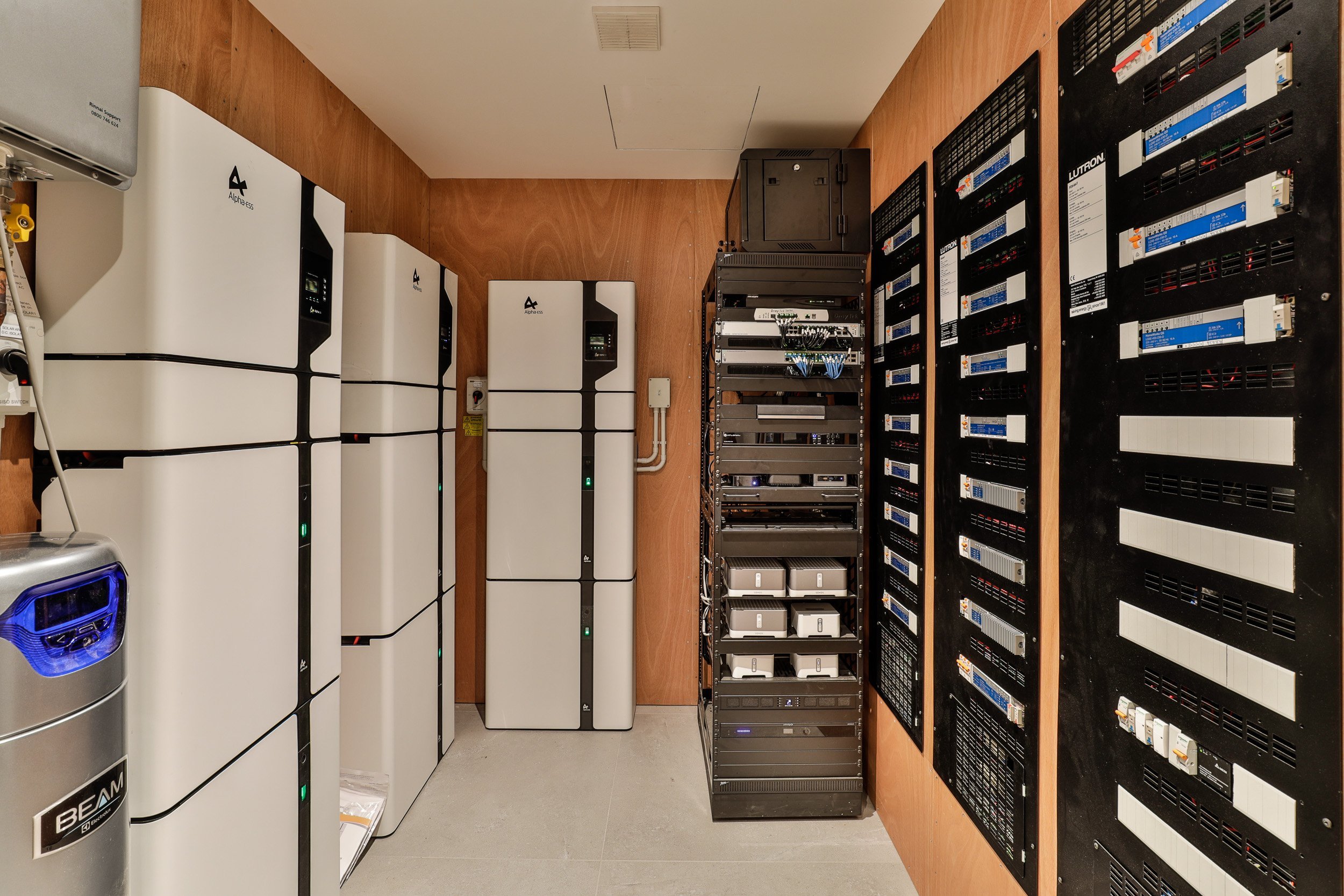 A tech room featuring white energy storage units, electronic equipment shelves, and wiring on wooden walls, emphasizing modern infrastructure.