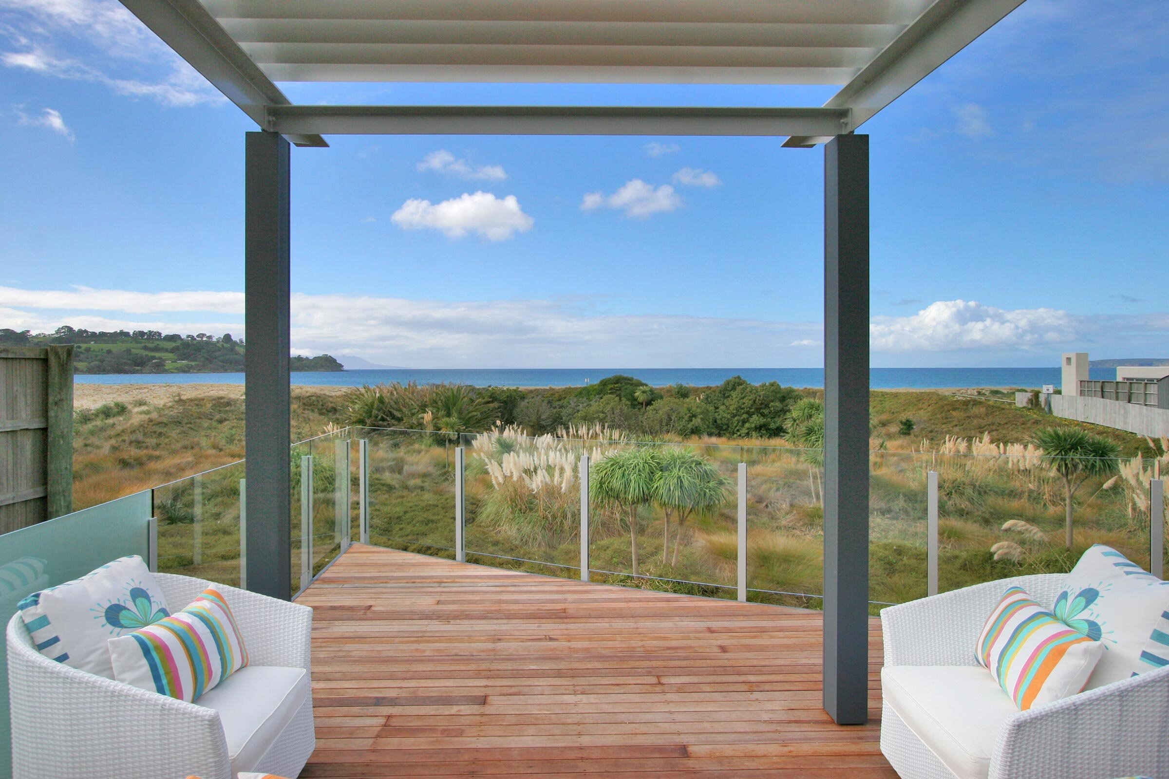 A cozy outdoor deck with white chairs overlooks a sandy beach and lush greenery, under a blue sky dotted with clouds.