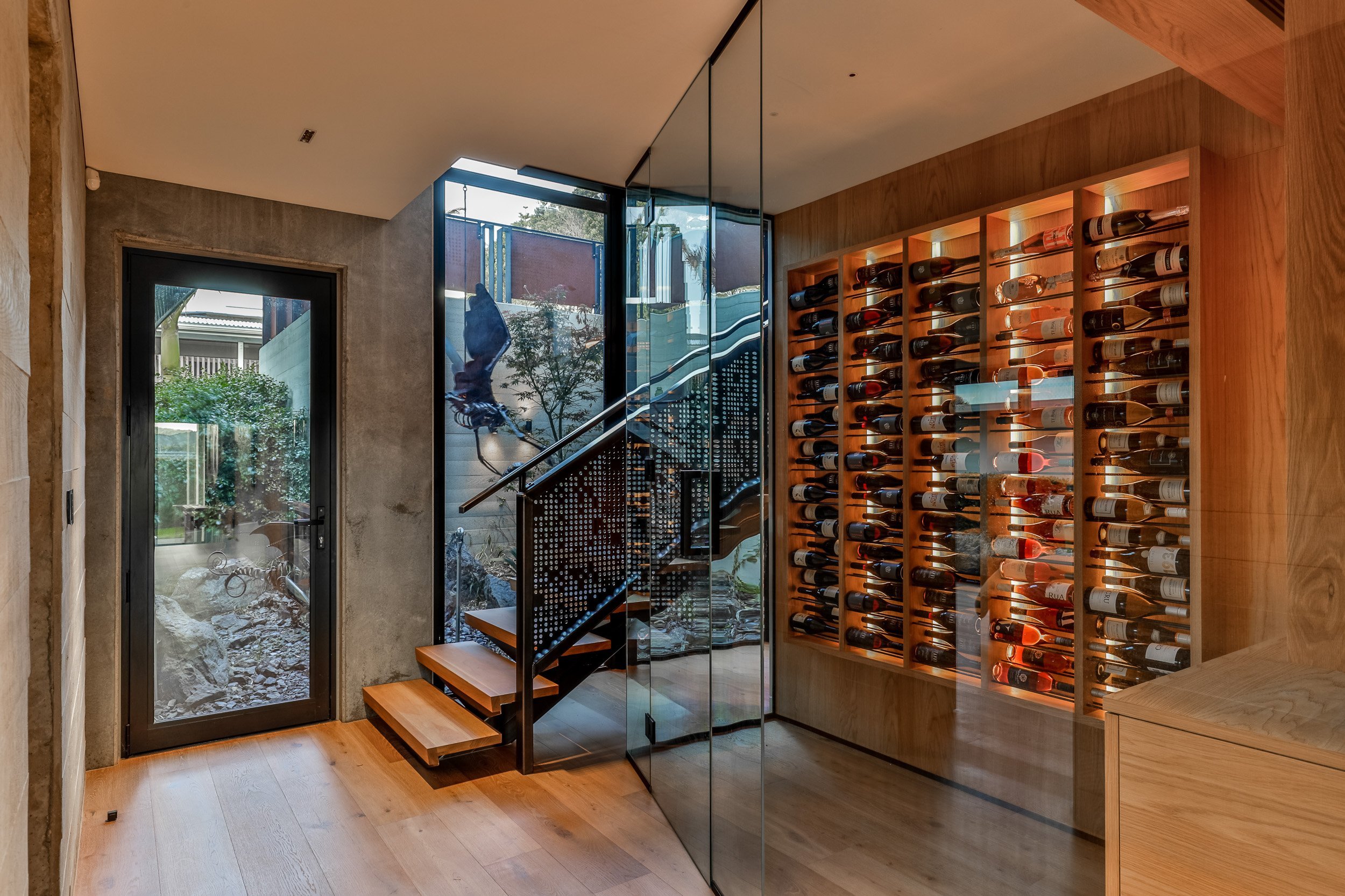 A modern wine cellar featuring a wall of illuminated wine bottles, glass walls, and wooden stairs leading to another level.