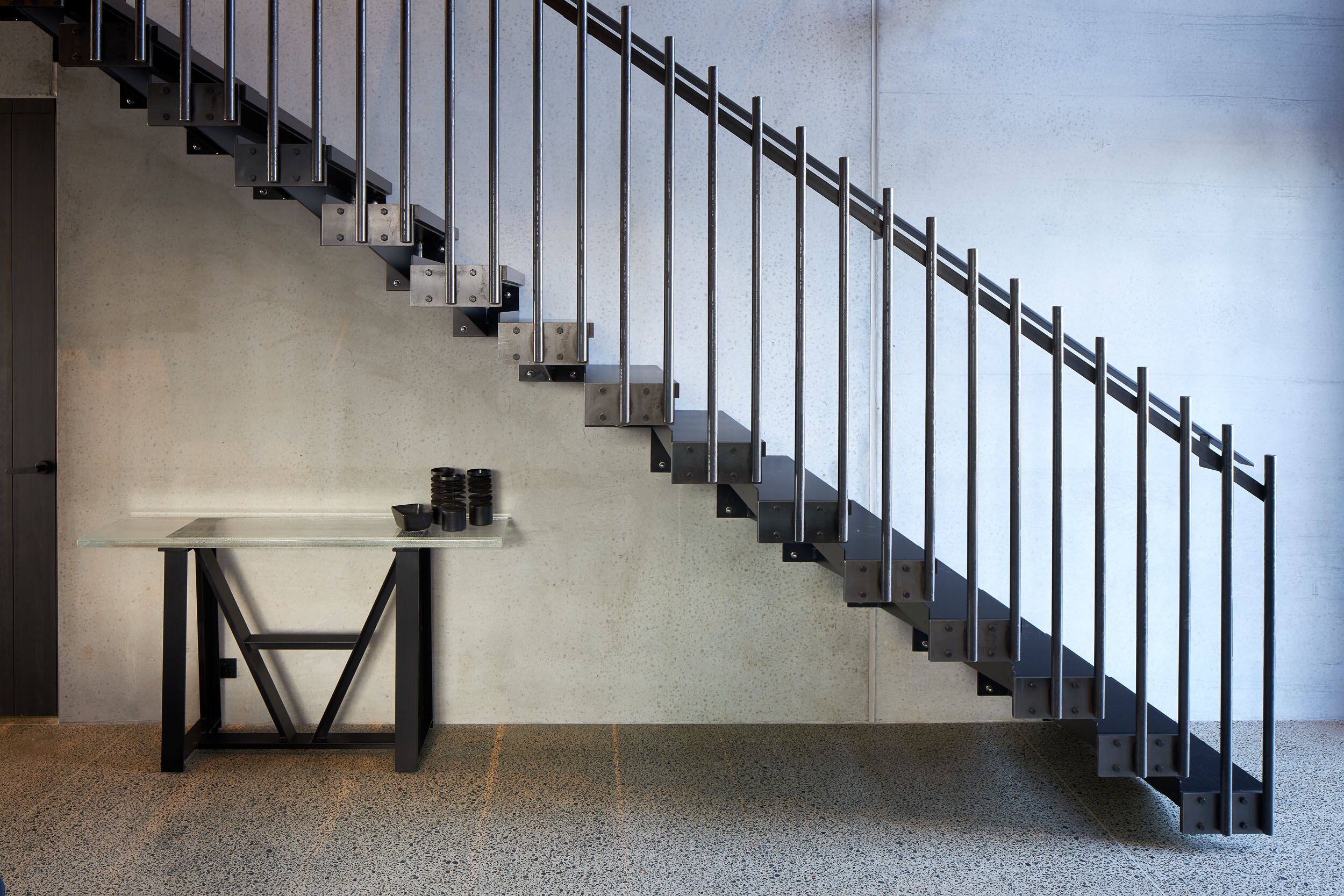 Modern staircase with metal railings and glass table below, set against a sleek concrete wall. Minimalist interior design focus.