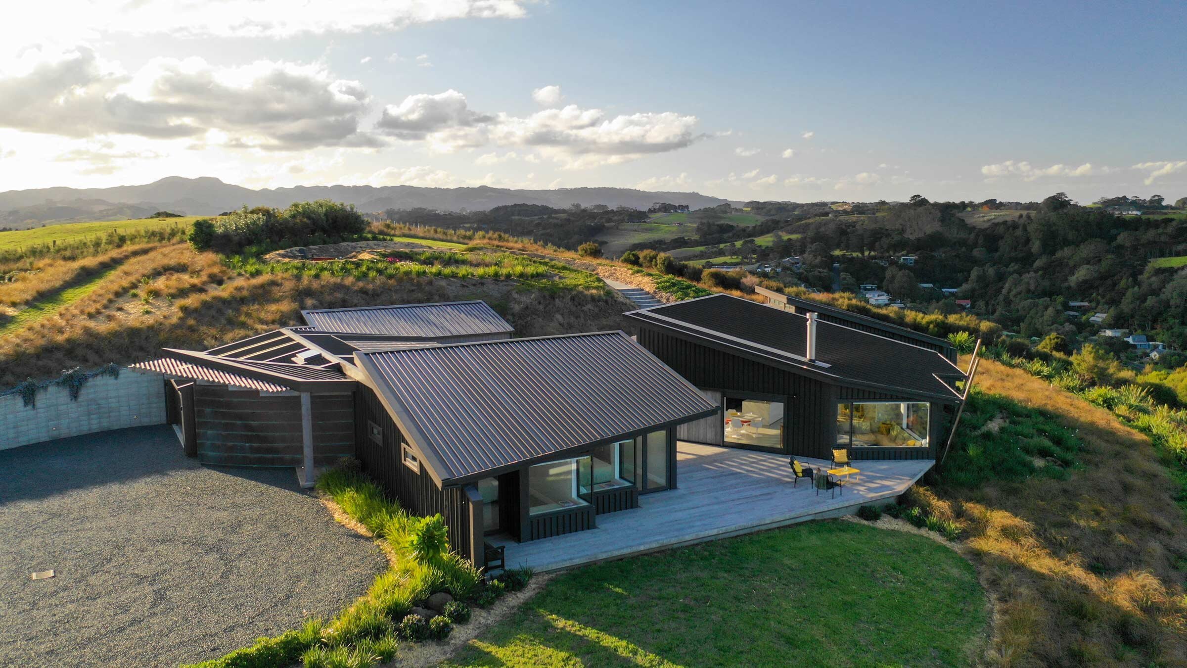 A modern black house with large windows is nestled on a hillside, surrounded by green fields and rolling hills under a blue sky.