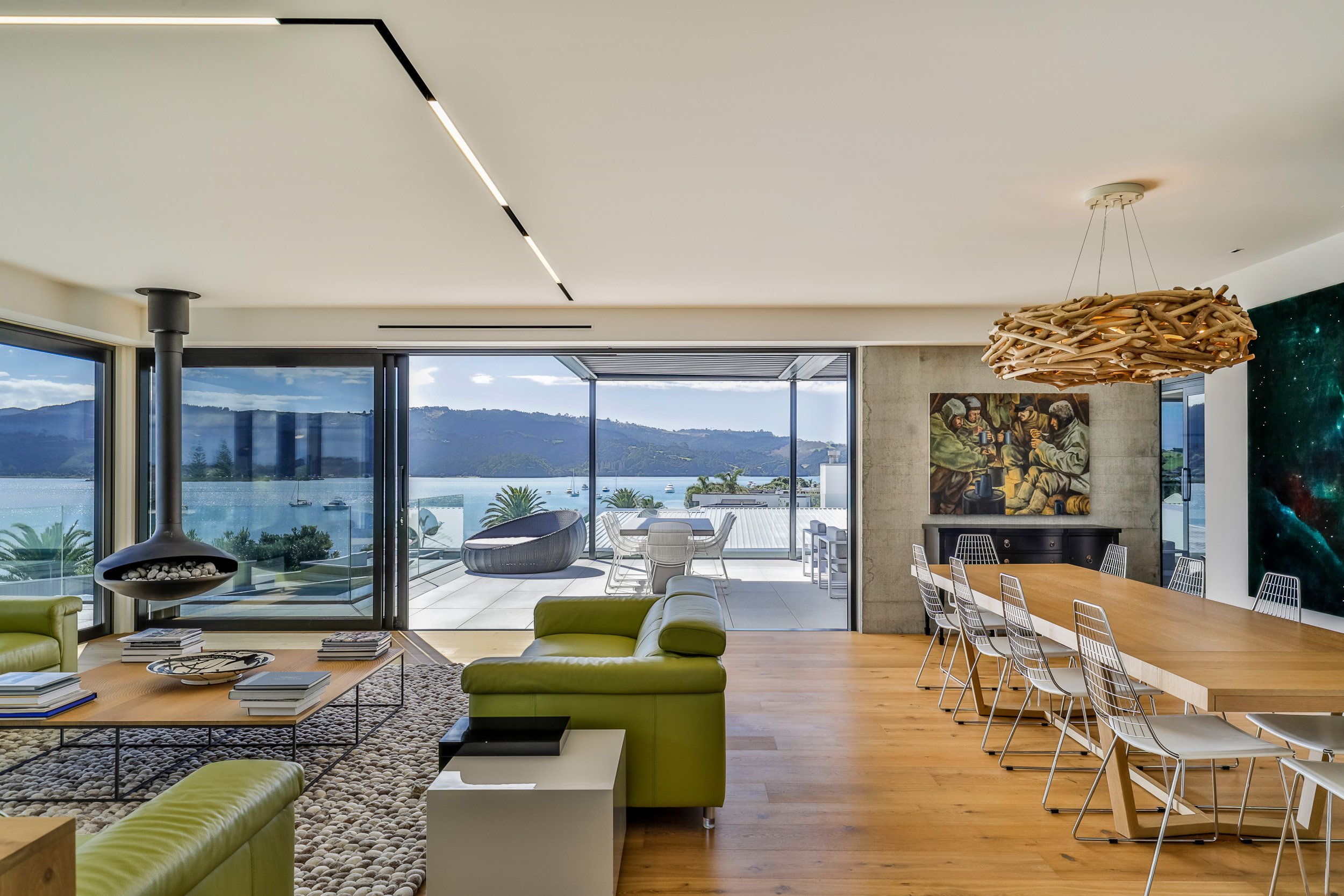 Modern living space with large windows overlooking a tranquil bay, featuring stylish green sofas and a unique fireplace.