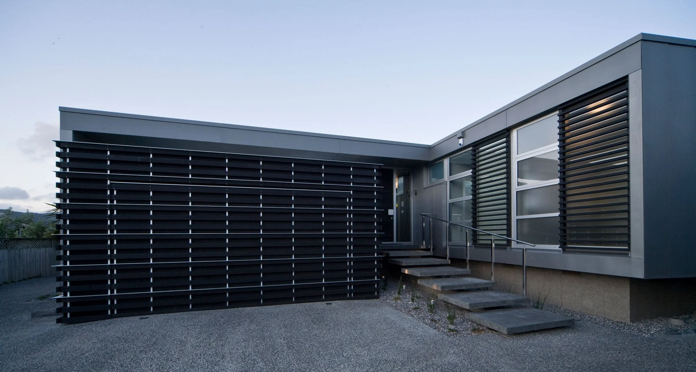 Modern residential building with a sleek design, featuring horizontal slatted black panels and large windows, approached by stone steps.
