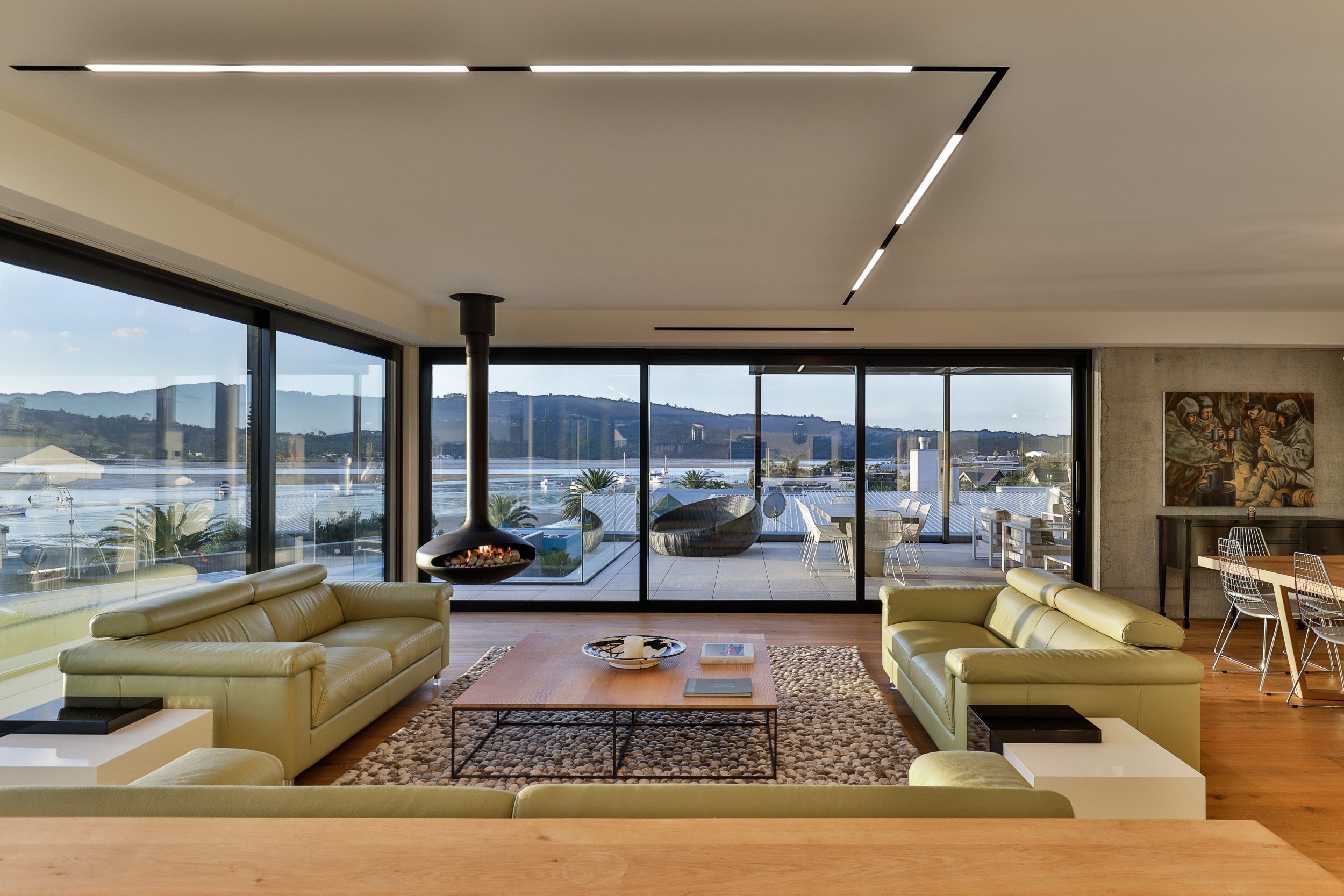 Modern living space with large windows overlooking a tranquil bay, featuring stylish green sofas and a unique fireplace.