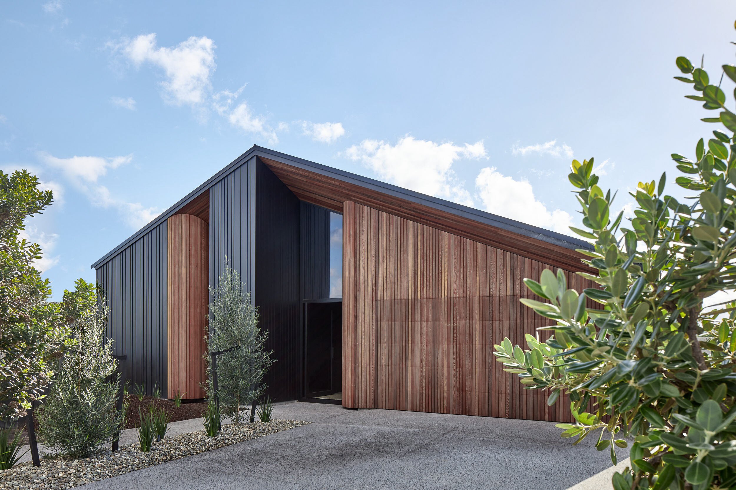 Modern house with a sleek black and wood exterior, surrounded by greenery and a clear blue sky.