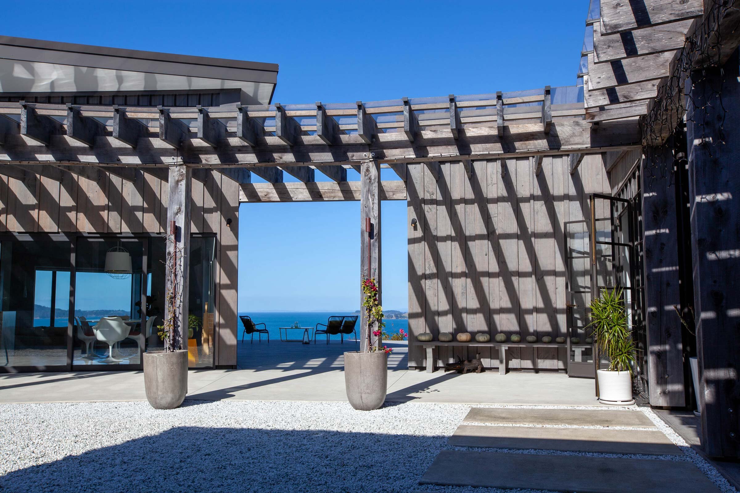 Contemporary coastal home entrance with a wooden pergola, leading to a patio with ocean views, surrounded by gravel and greenery.