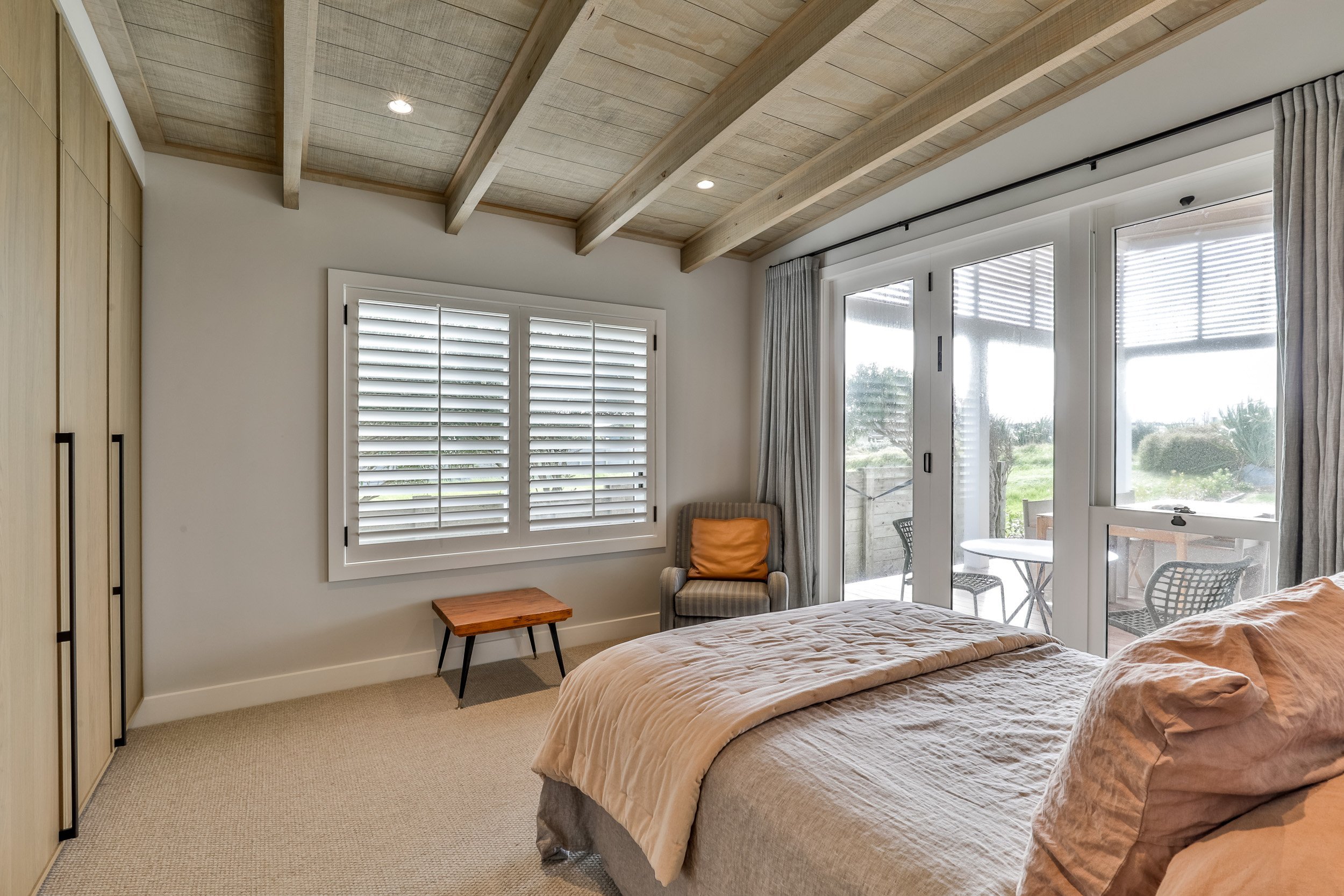 Cozy bedroom featuring a neutral palette, wooden beams, large windows with blinds, and a small patio outside with garden views.