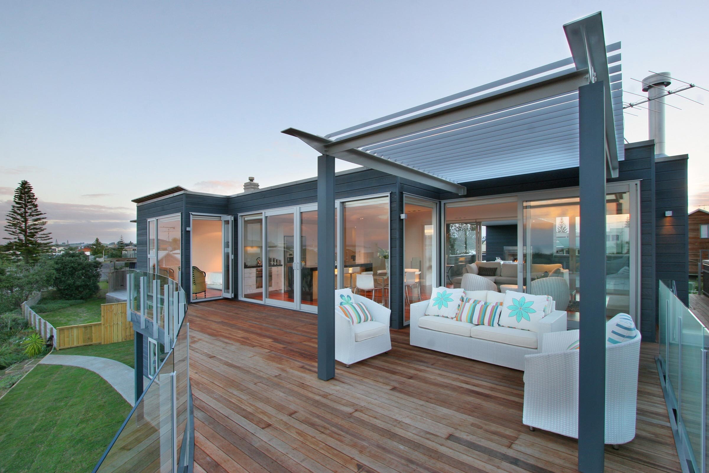 Modern house with a wooden deck, glass railings, and stylish outdoor seating, surrounded by greenery and a tranquil evening sky.
