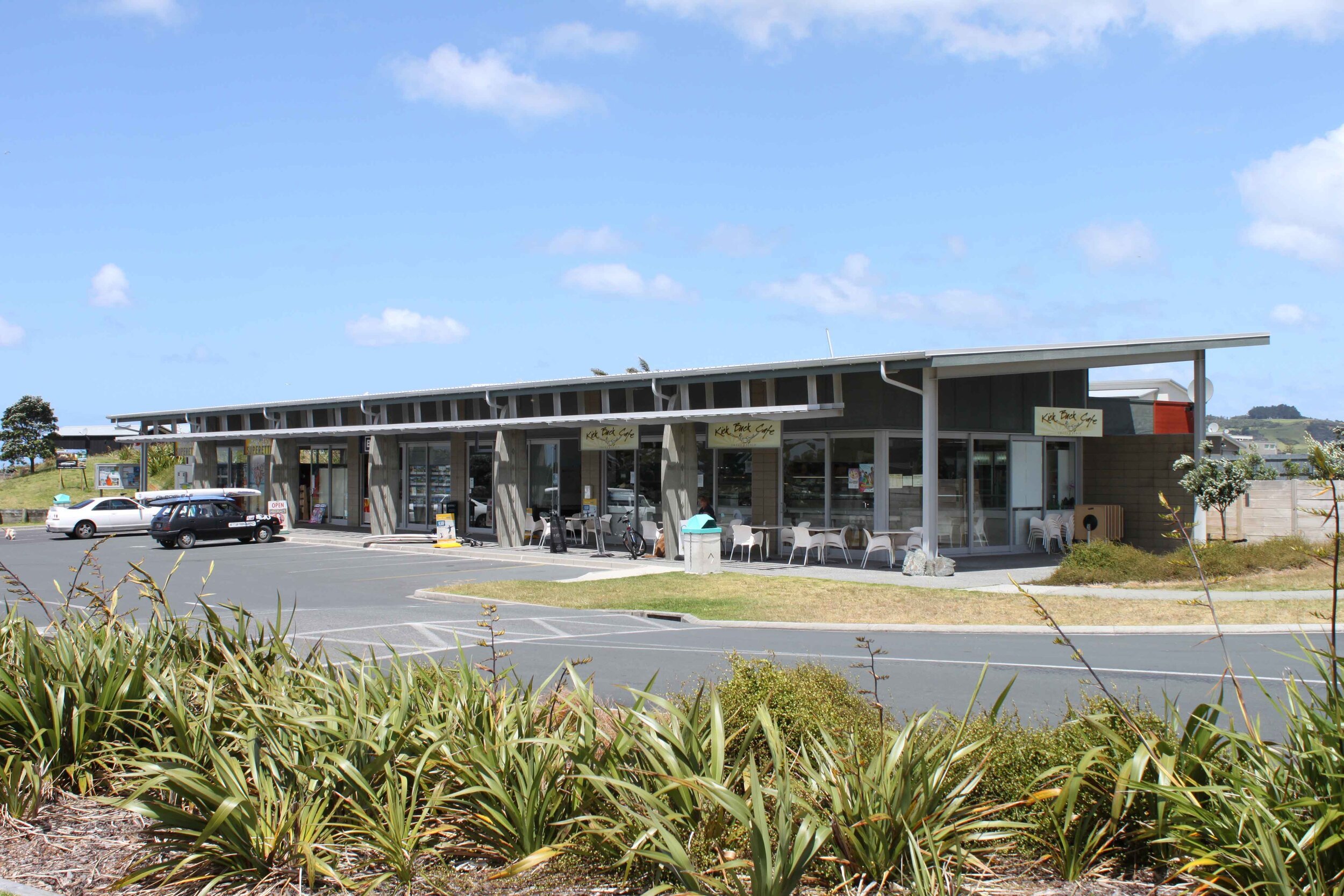 A modern cafe building with large windows, outdoor seating, and greenery in front, set against a blue sky and distant hills.