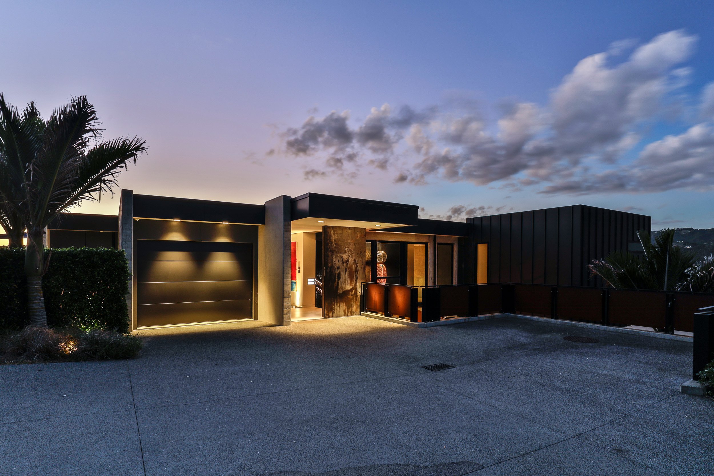 Modern house exterior at dusk, highlighting a sleek design with warm lighting, a garage, and surrounding palm trees.