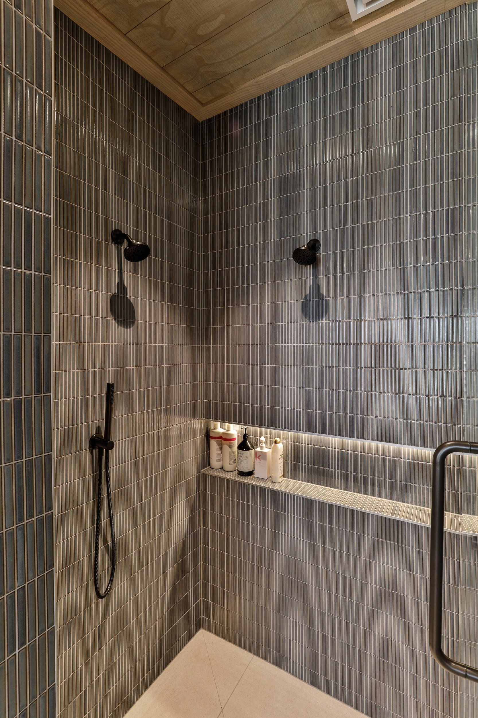 Modern shower with gray tiled walls, two black showerheads, a sleek shelf with various bottles, and a wooden ceiling.