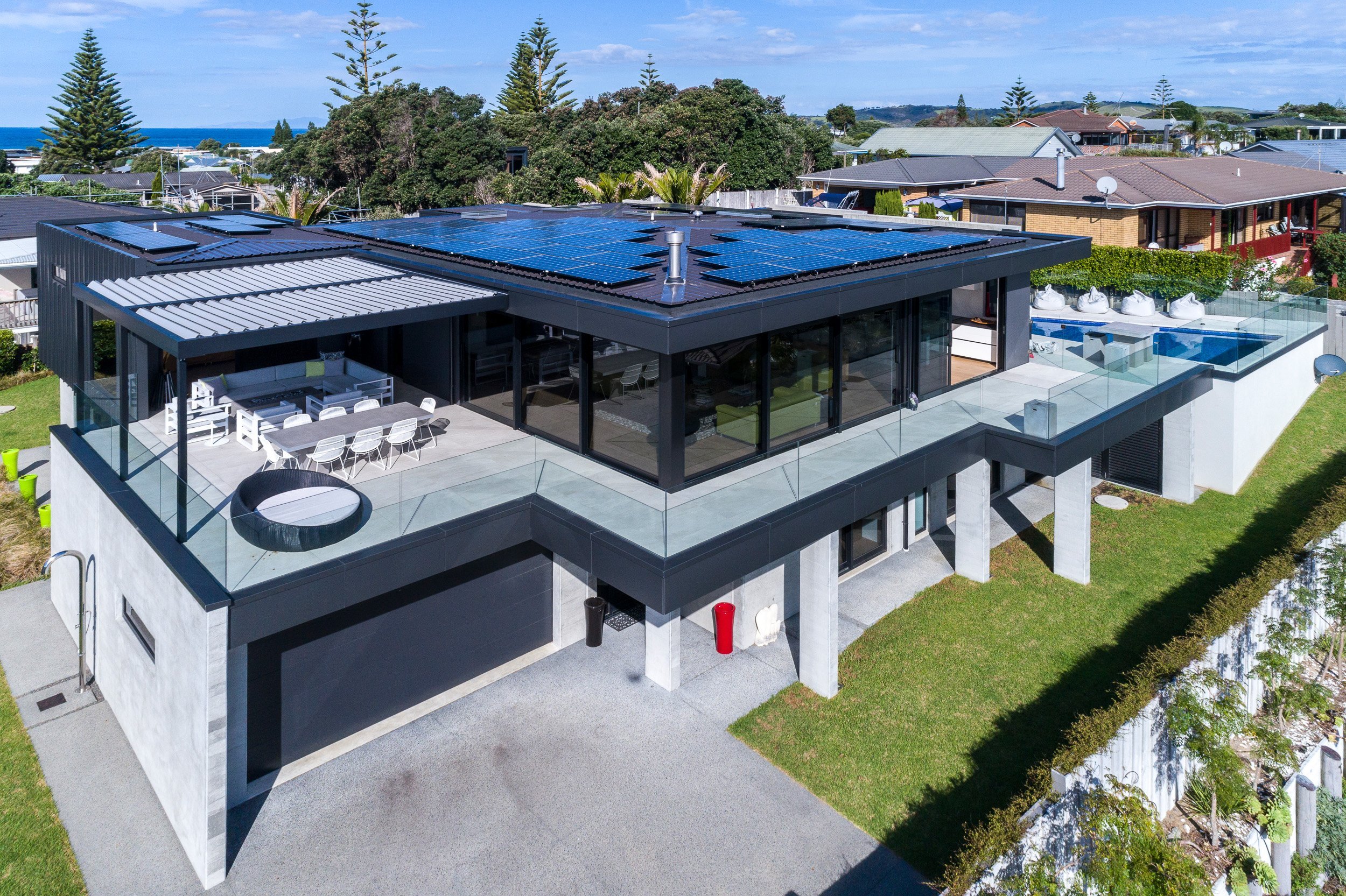Modern two-story house with solar panels, outdoor seating area, pool, and landscaped yard, overlooking the ocean in a sunny locale.