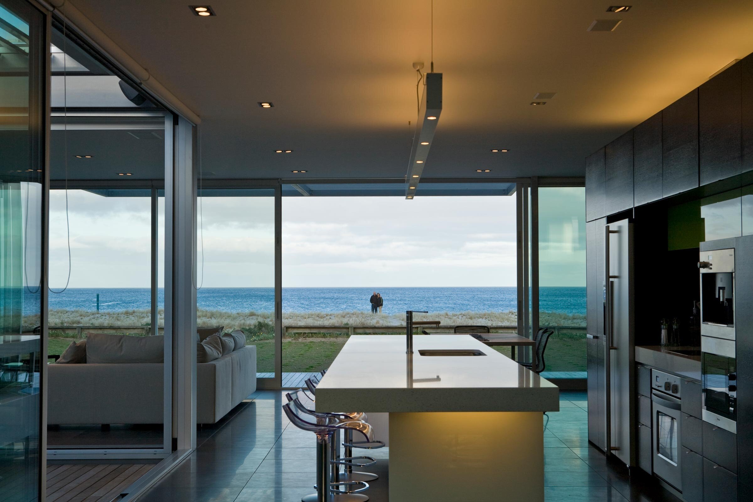 Modern kitchen with a sleek countertop overlooking the ocean, framed by floor-to-ceiling windows and a cozy living area behind.