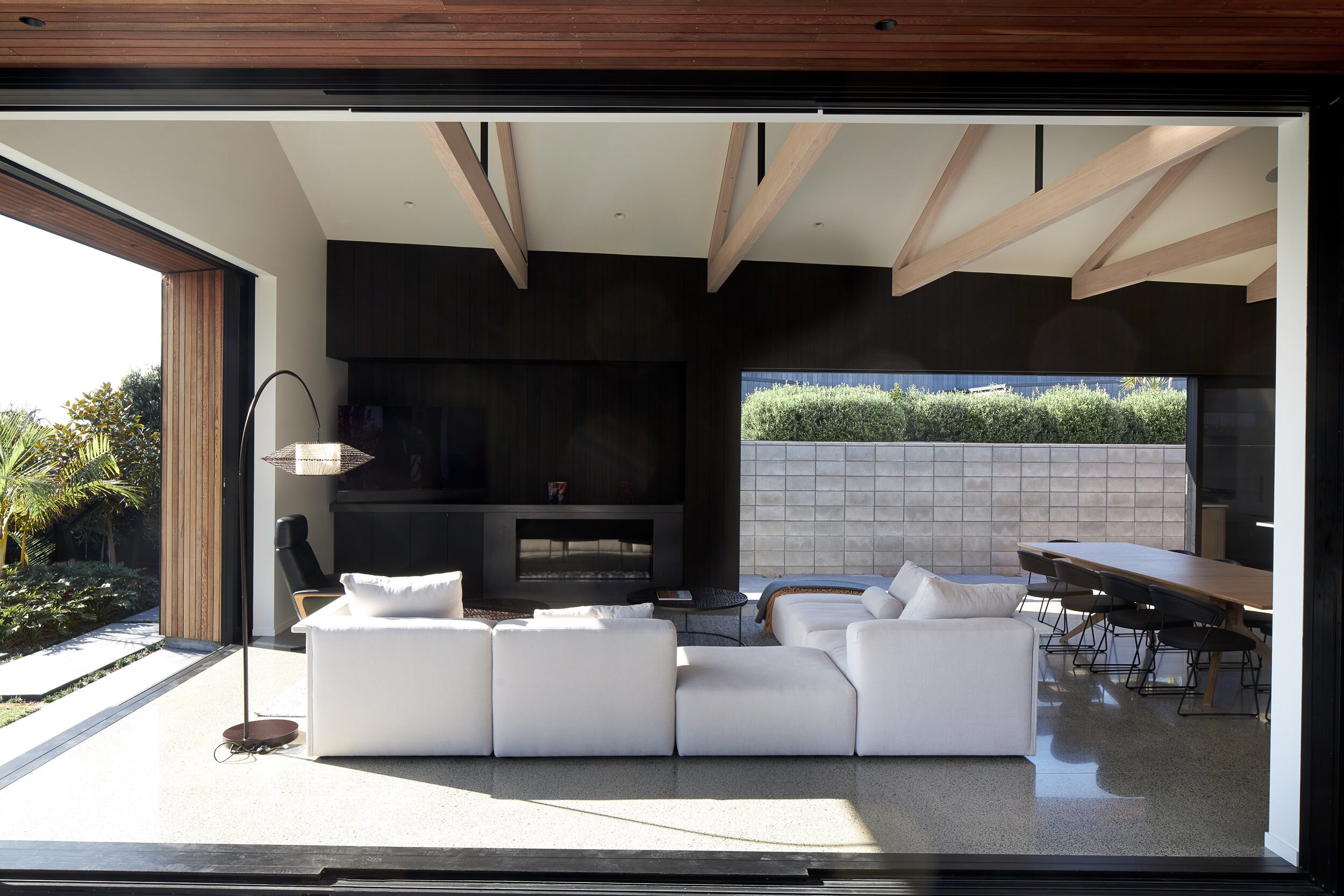 Modern living room with white sectional sofa, wooden beams, and large opening to a patio, featuring a dining table and greenery outside.