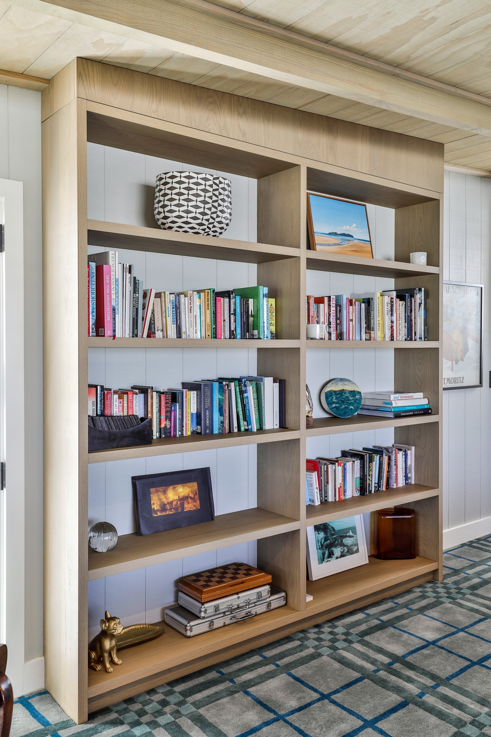 A modern wooden bookshelf filled with colorful books, decorative objects, and framed photos, against a light wall and patterned carpet.