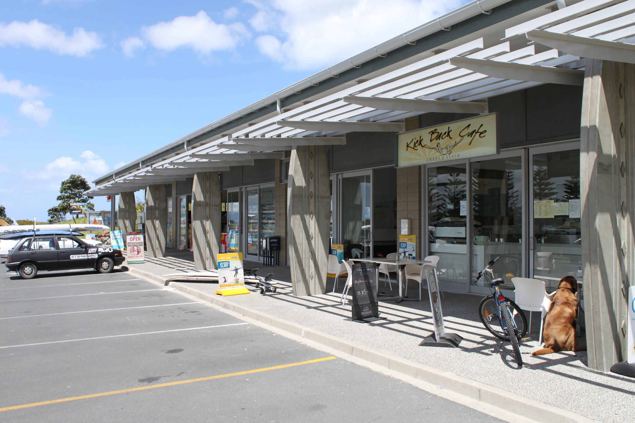 A sunny café exterior with outdoor seating, a parked car, a bicycle, and a dog resting near the entrance. "Kick Back Café" sign visible.