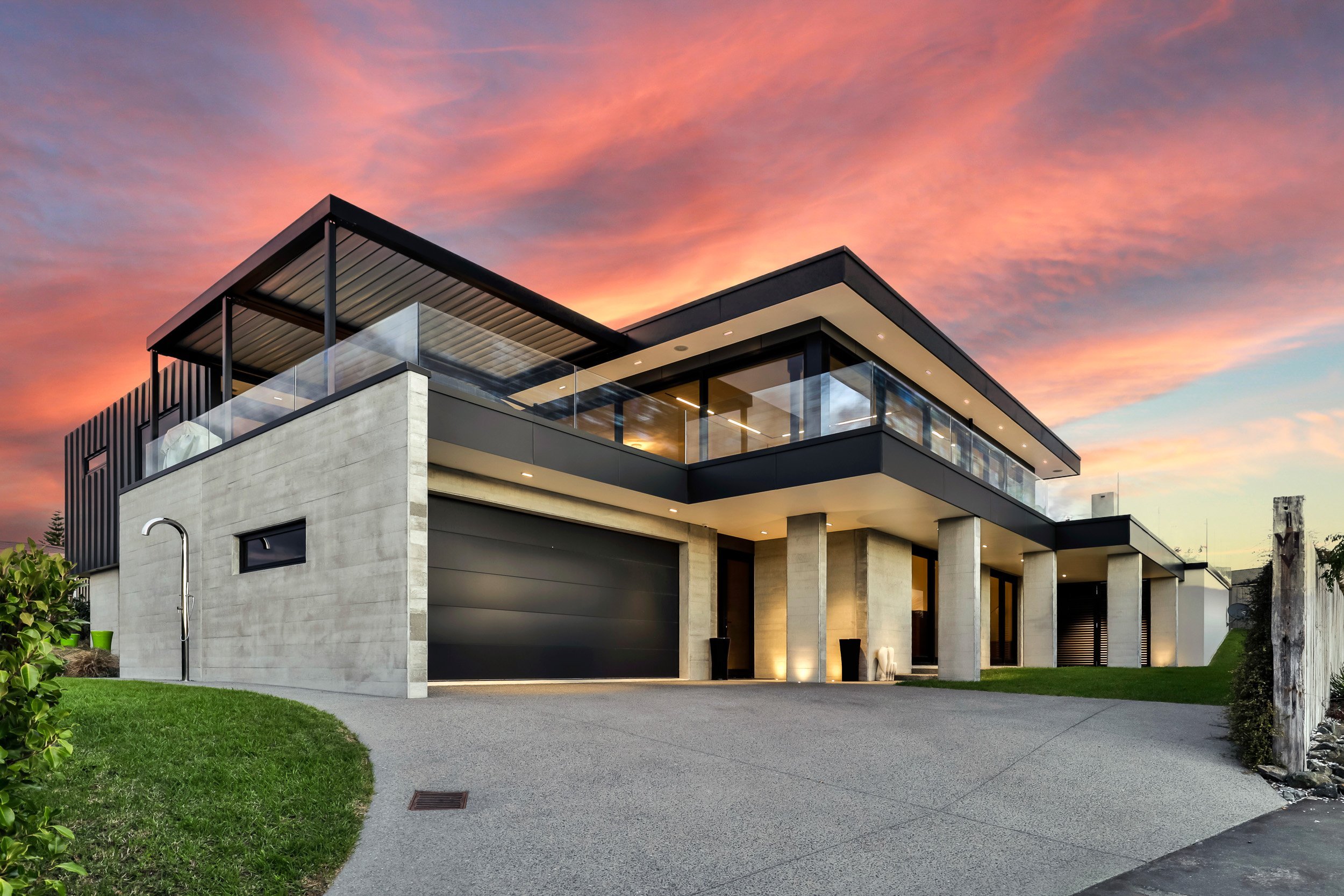 Modern two-story house with a sleek design, glass balcony, and dramatic sunset sky, featuring landscaped surroundings and a unique driveway.