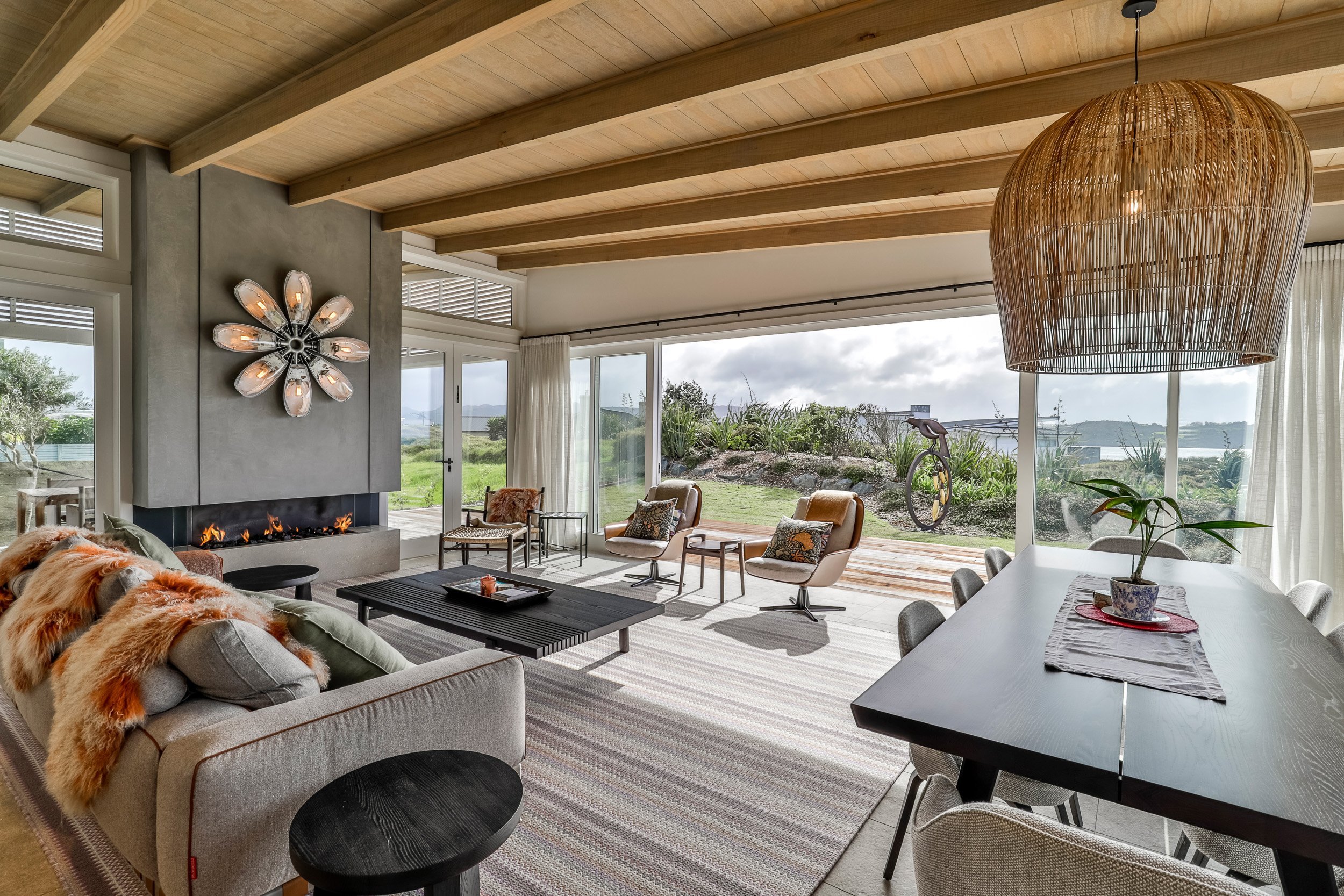 Modern living room with wooden beams, cozy furniture, and a fireplace. Large windows offer scenic outdoor views.