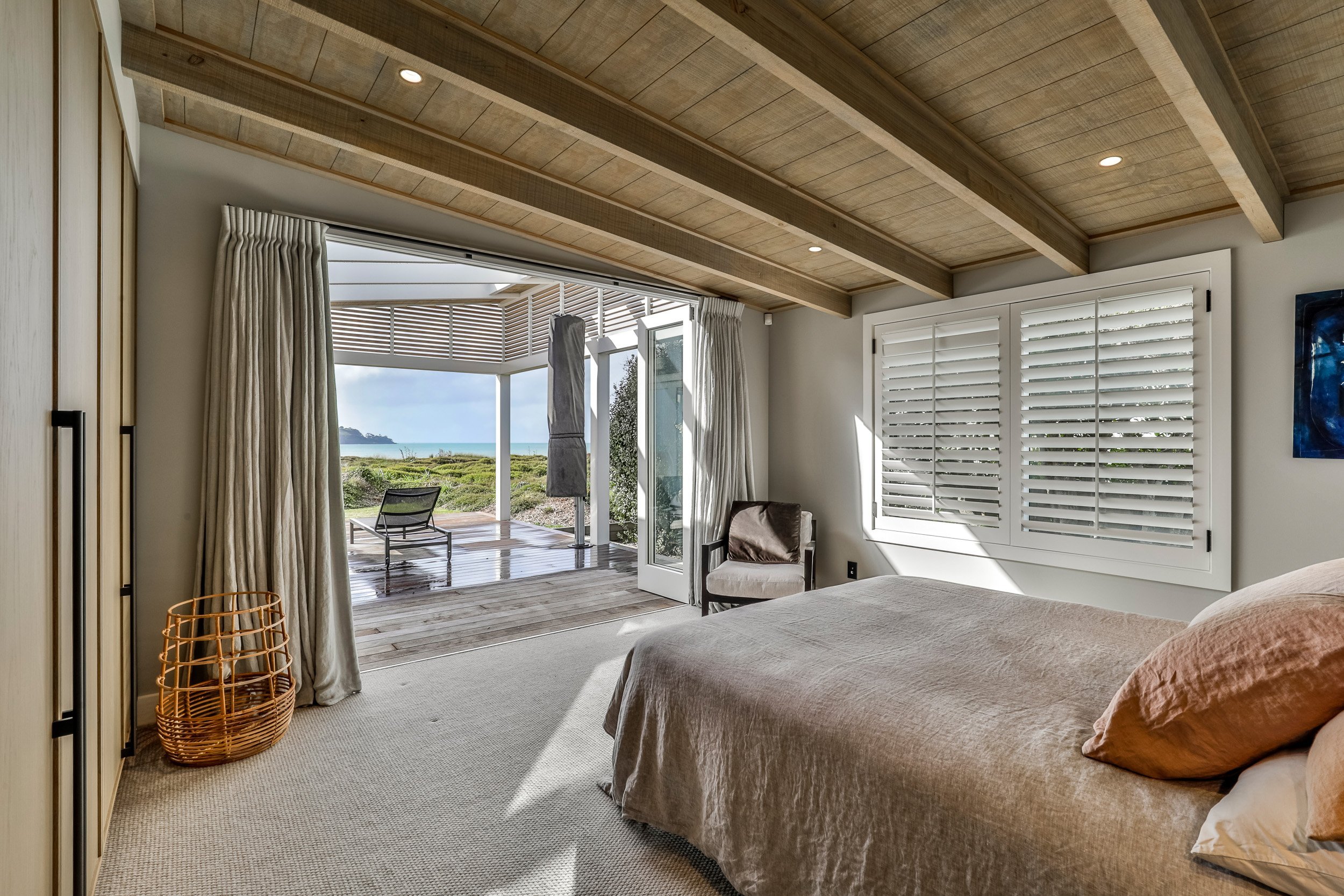 Cozy bedroom with a light-colored quilt, wooden beams, modern furniture, and a view of outdoor seating through glass doors.