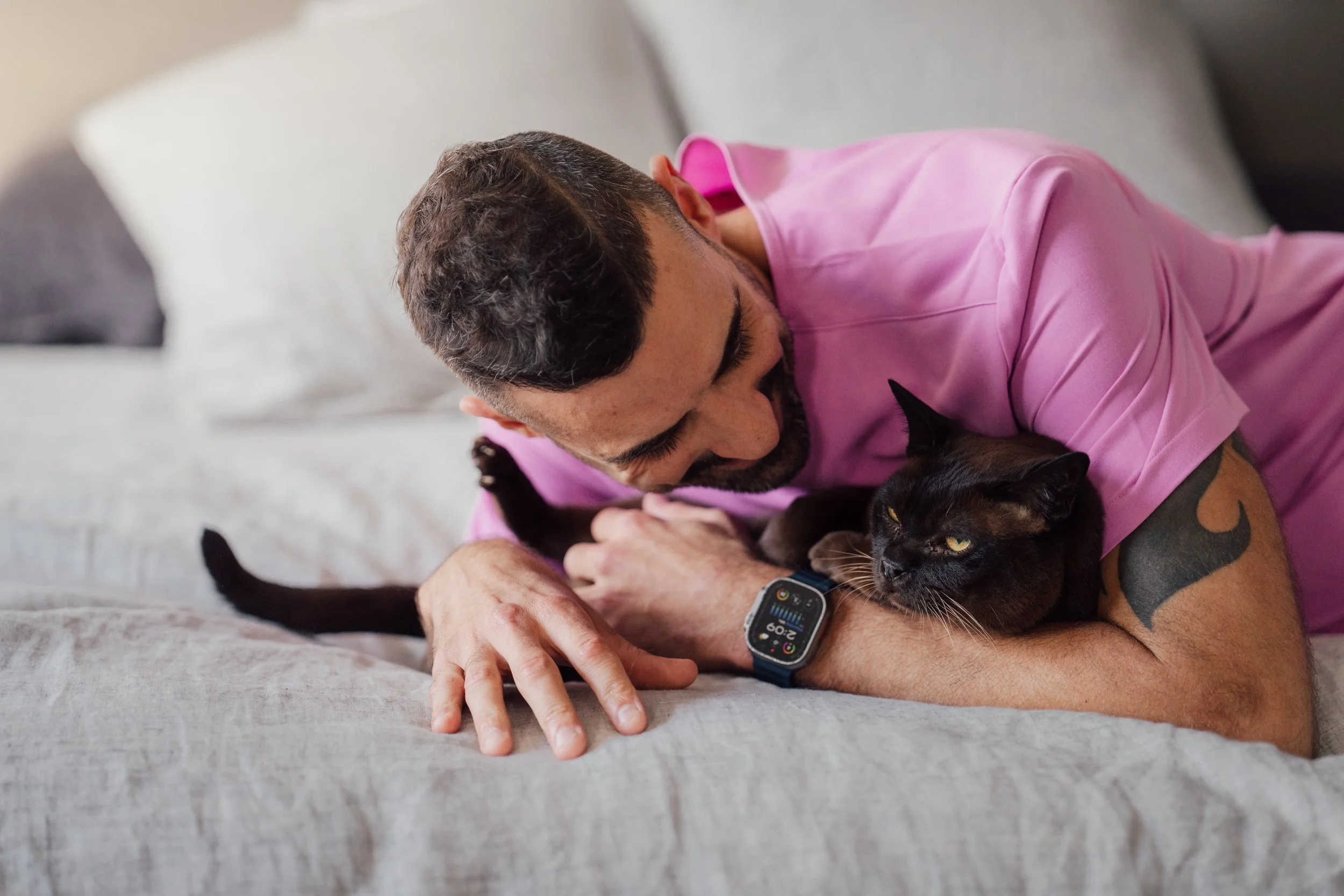 A man with a beard and tattoo on his arm lying on a bed, wearing a pink shirt, playing with a black cat that is lying on his arm.
