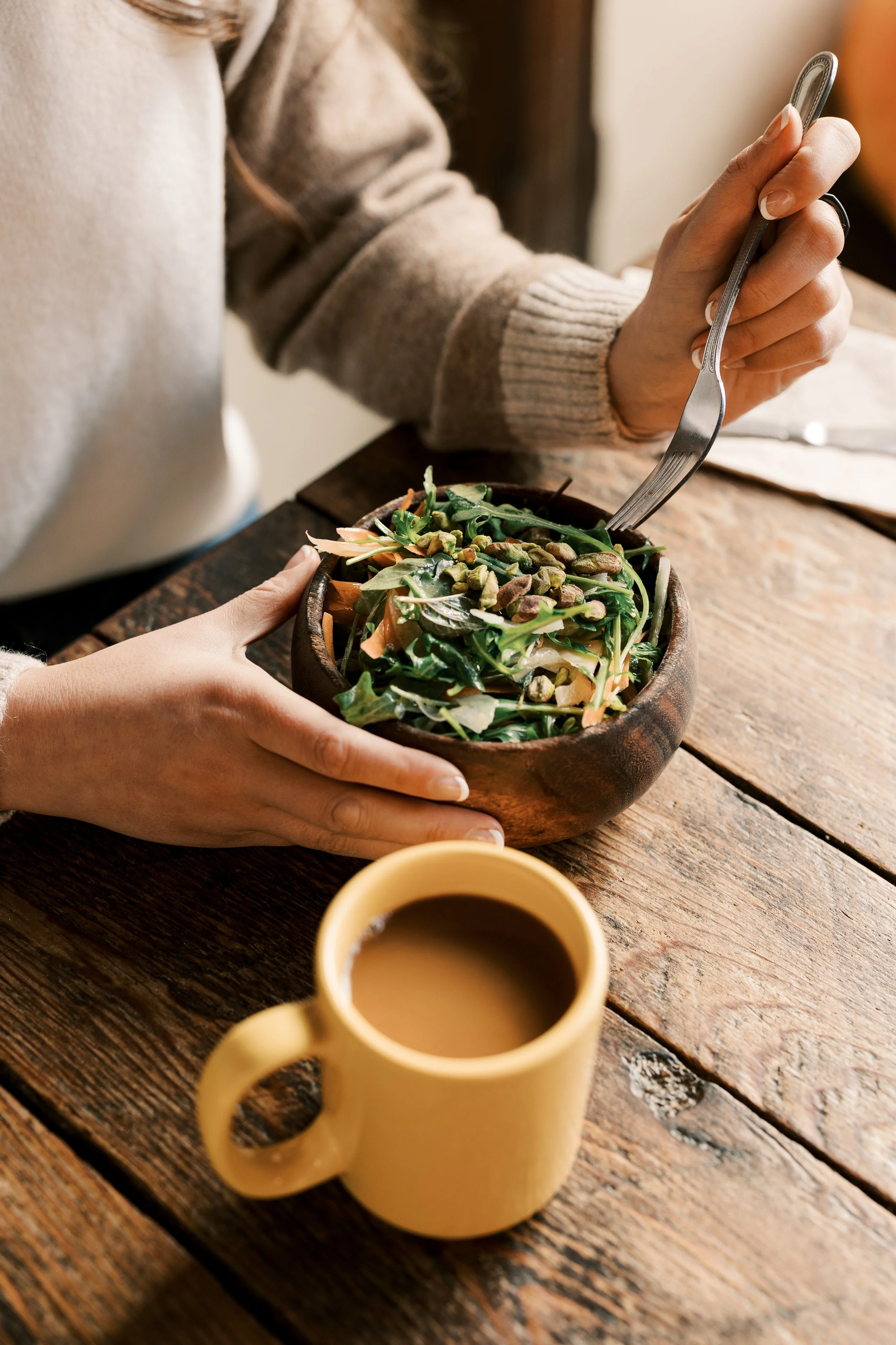 Person holding a salad bowl with a fork, sitting at a wooden table with a yellow mug of coffee nearby.