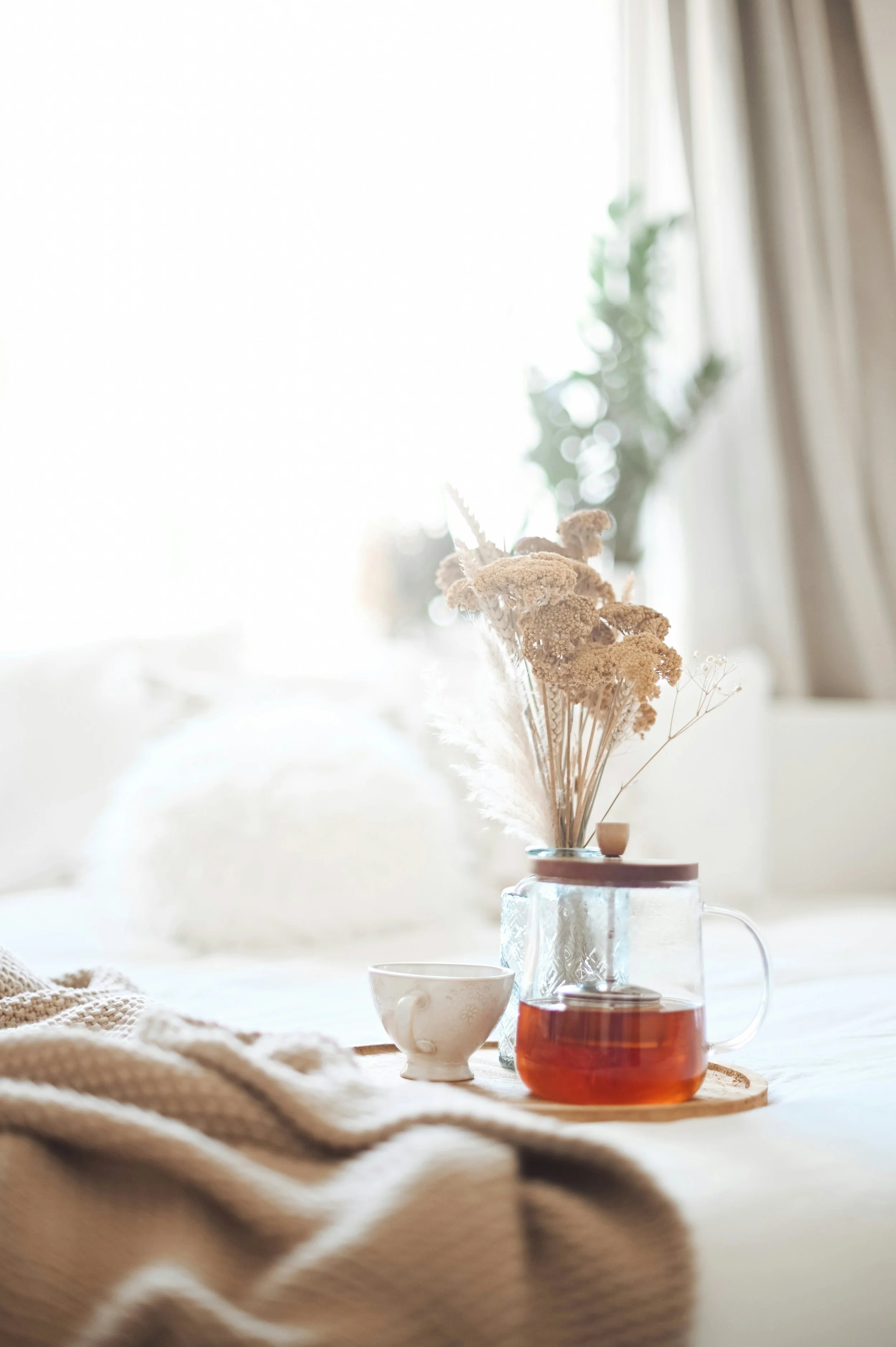 A cozy indoor scene with a teapot filled with tea, a white teacup, a vase with dried flowers, and a textured blanket on a table, with sunlight streaming in through a window.