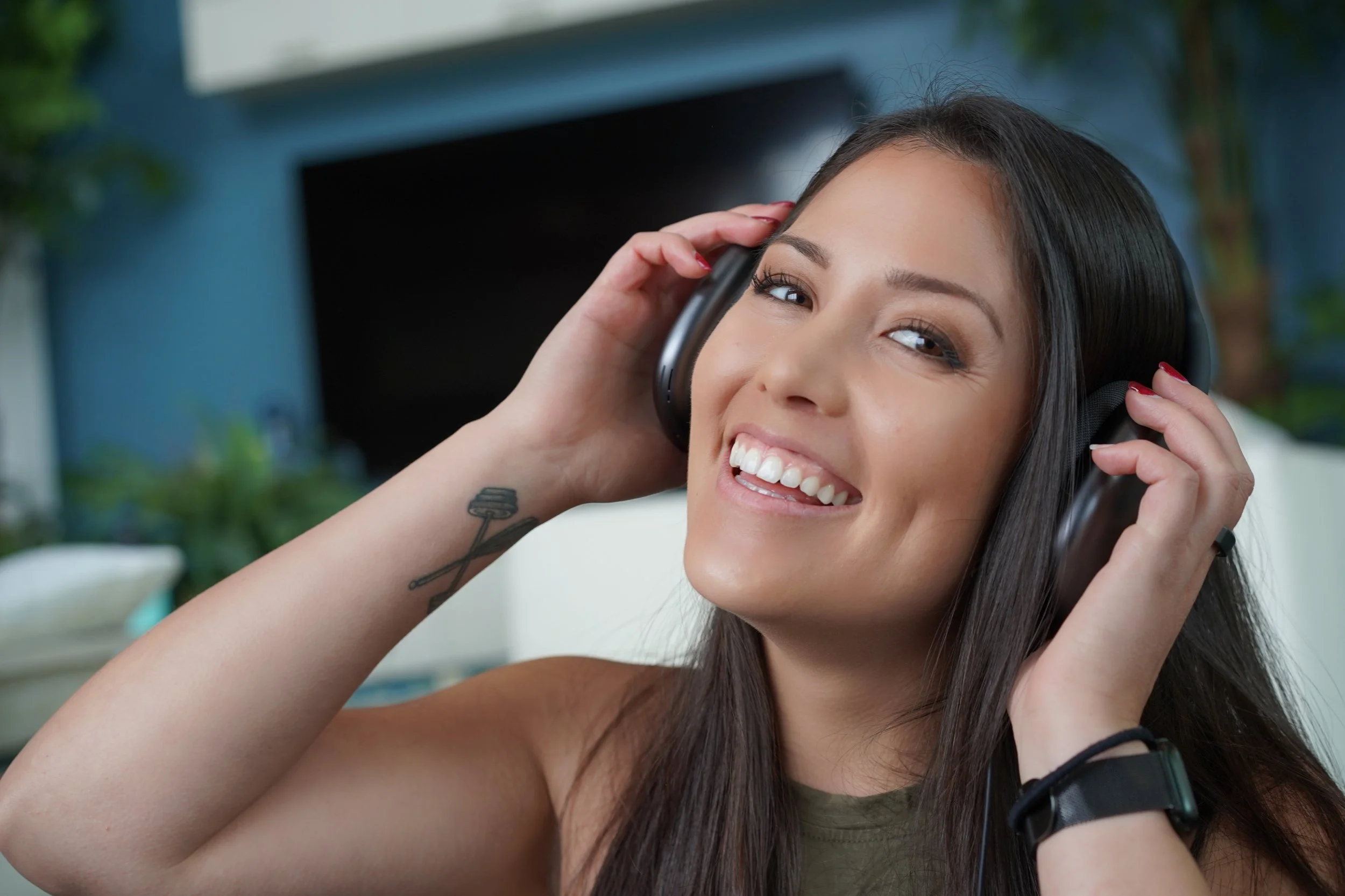 A young woman with long dark hair smiling happily while wearing headphones, in a living room with a blue wall and a television in the background.