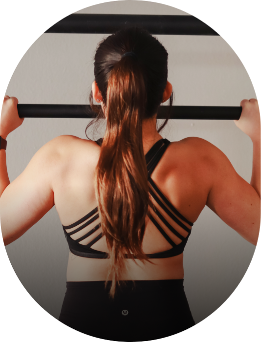 Coach Kailes performing a pull up exercise with a bar, back view, wearing a black sports bra with strap details.