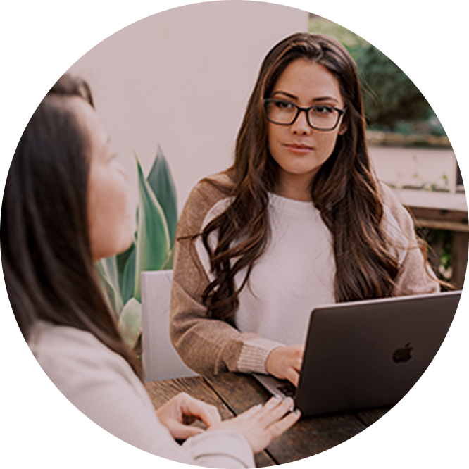 Two women having a conversation at a table, one with glasses and long dark hair, the other with shoulder-length hair, and a laptop in front of her