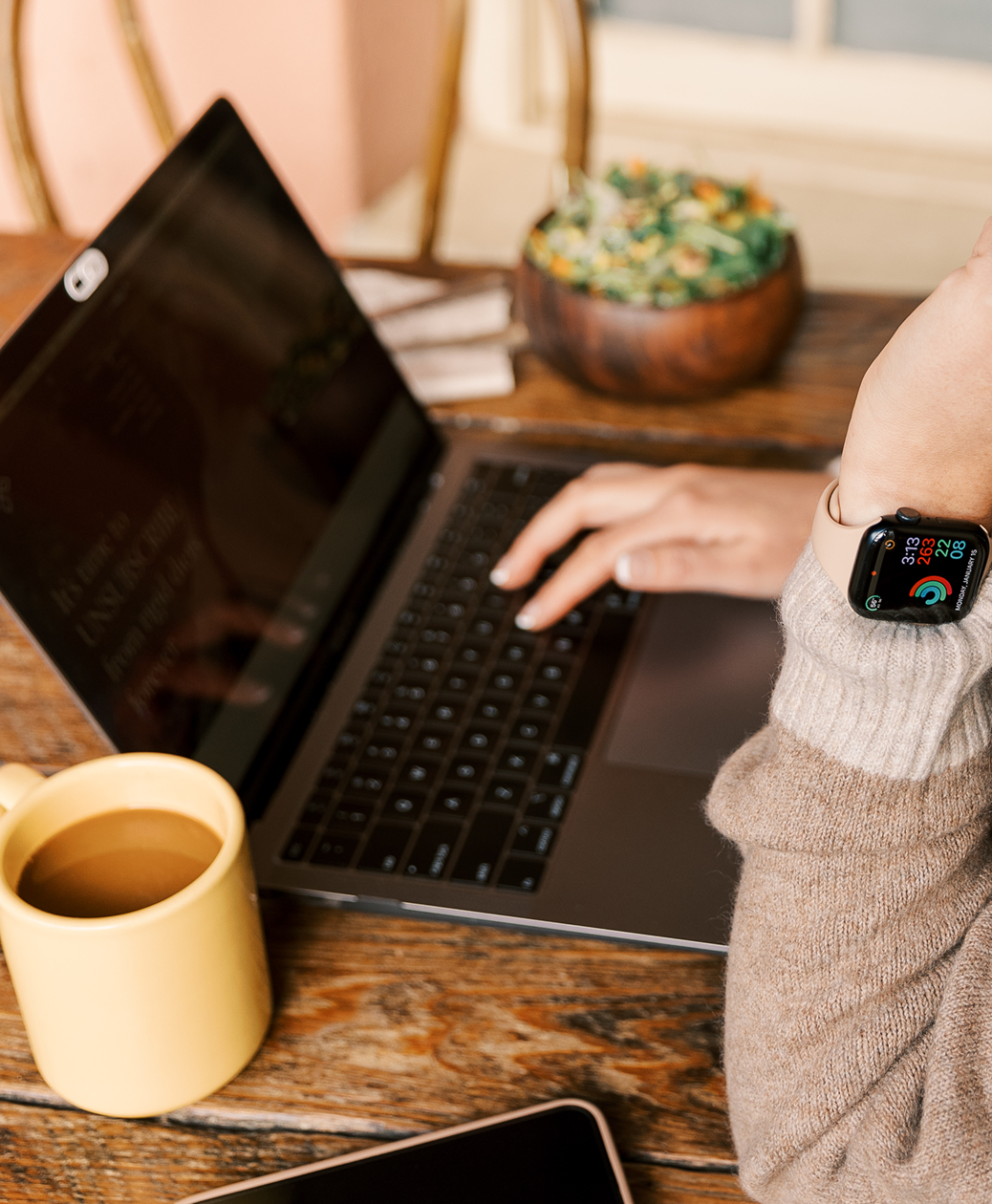 A person wearing a beige sweater and smartwatch working on a laptop at a wooden table with a yellow mug of tea, a bowl of salad, and a smartphone working on custom nutrition programming.