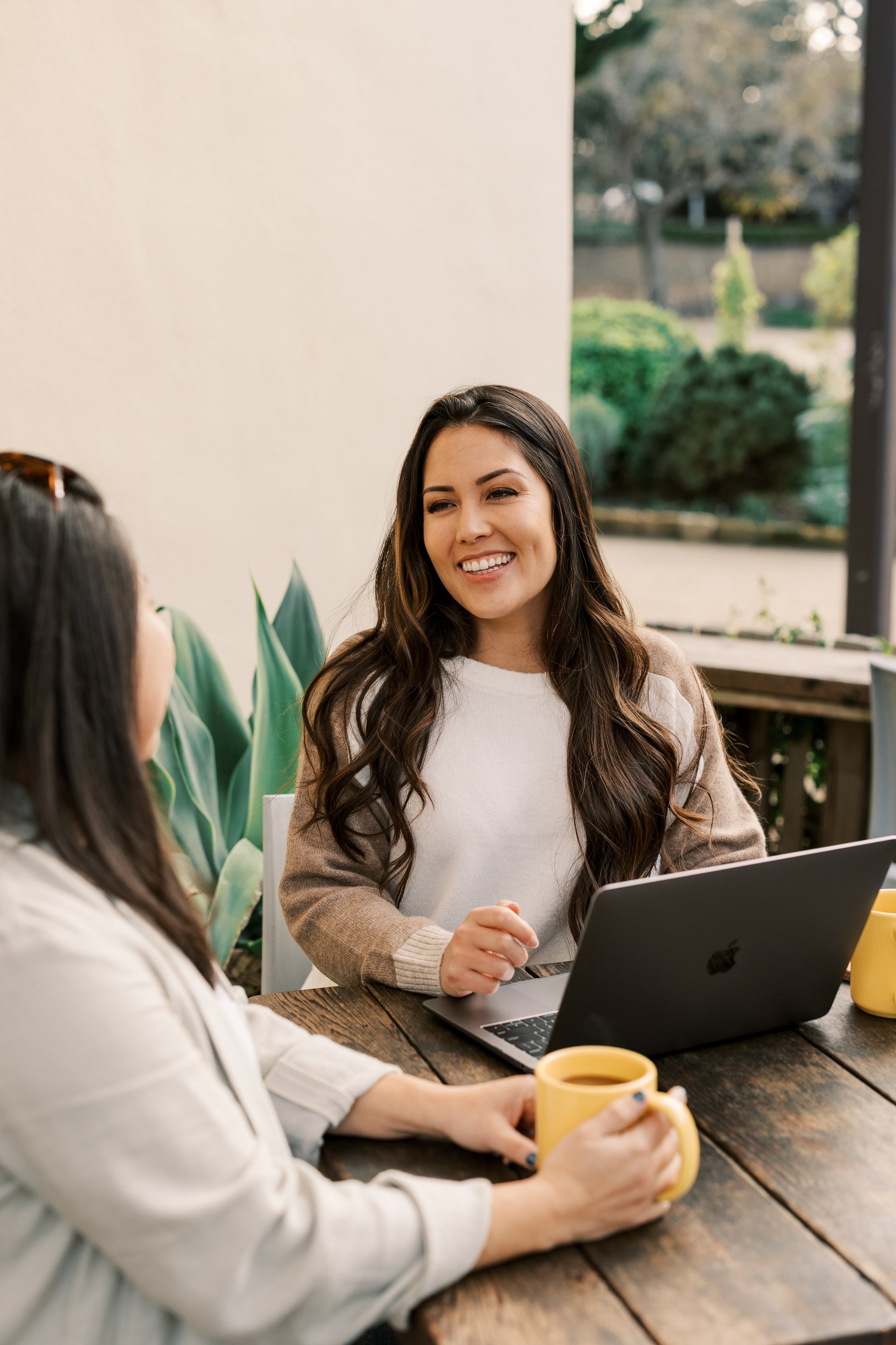 Two women sitting at a wooden table outdoors, engaged in conversation, with a laptop in front of them and yellow mugs in their hands, smiling and enjoying each other's company.