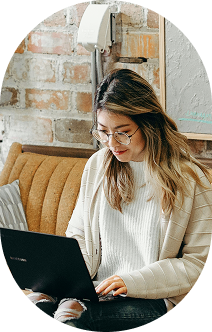 Young woman with glasses working on a black laptop in a cozy, modern space with a brick wall background and a brown cushioned seat.