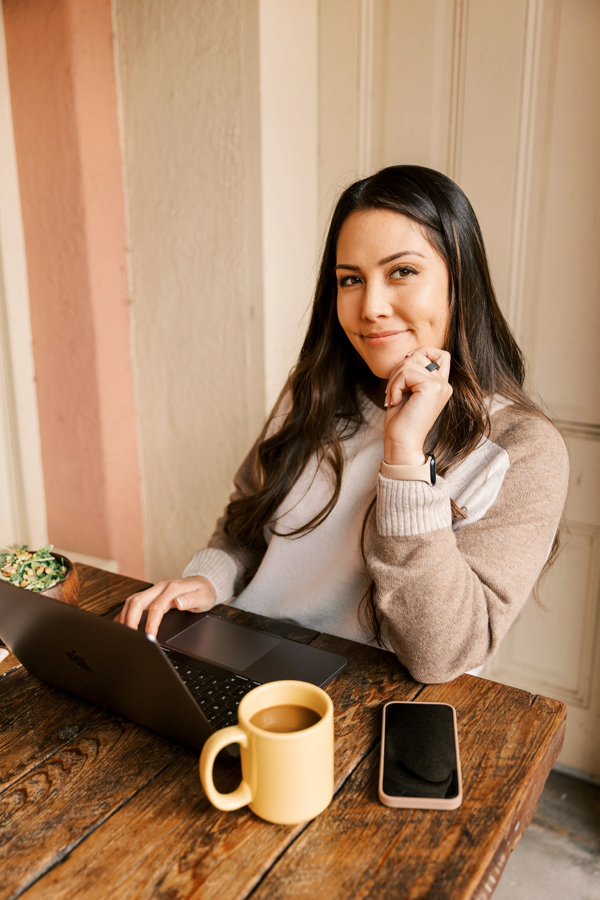 A woman sitting at a wooden table with a laptop, smartphone, and a yellow coffee mug. She has long dark hair, is wearing a beige and white sweater, and is smiling while looking at the camera.