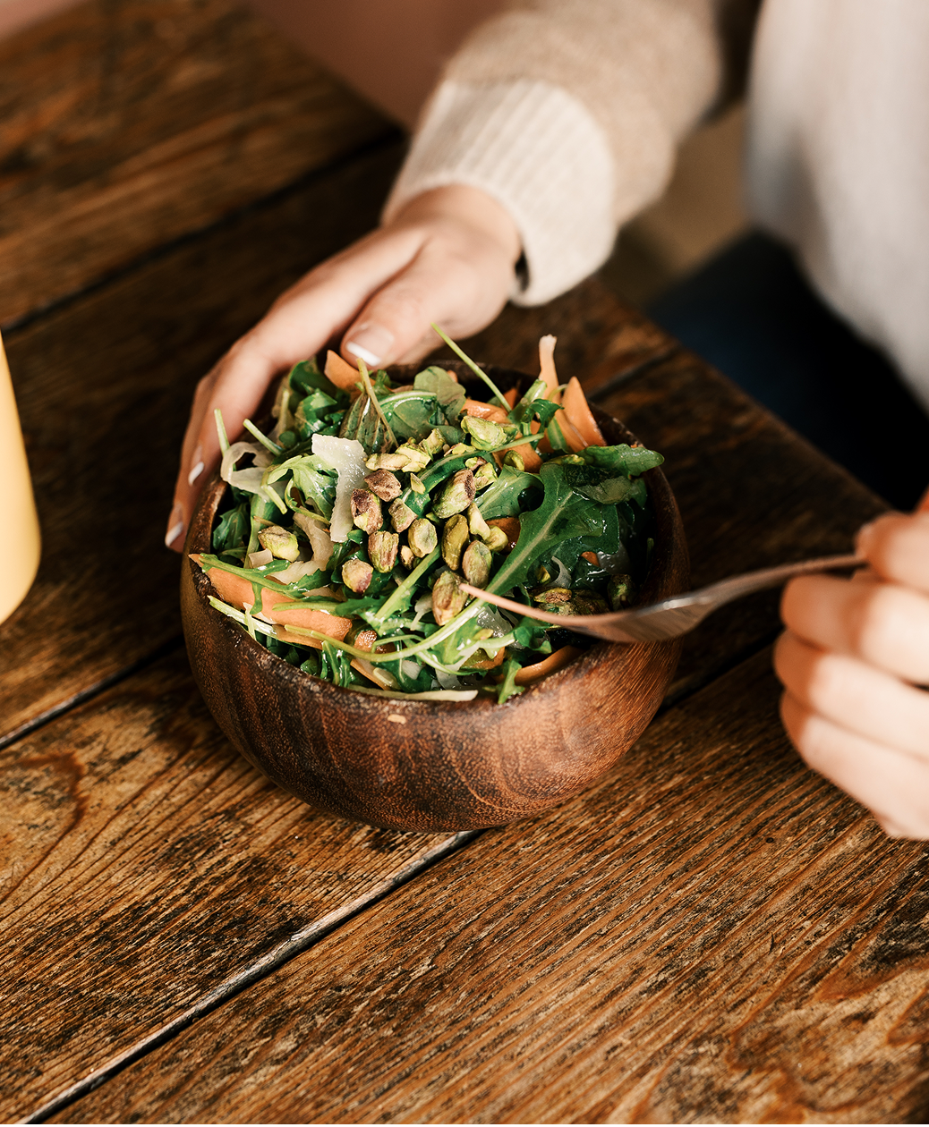 A person is holding a wooden bowl of salad with arugula, shredded carrots, shredded cheese, and pistachios, on a rustic wooden table.