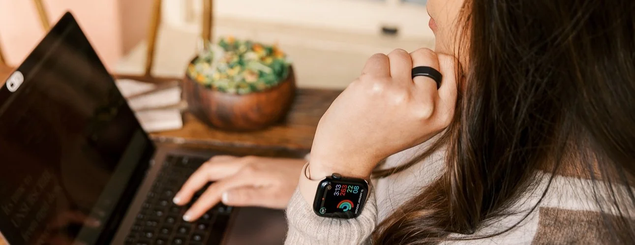 A person with long brown hair sitting at a desk, looking at a laptop screen while wearing a smartwatch and a ring, with a bowl of snacks in the background.