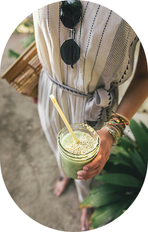 A woman holding a green smoothie in a clear glass with a yellow straw, wearing a striped dress, glasses hanging on her chest, and jewelry, standing outdoors near plants.