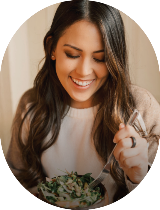 A woman with long brown hair smiling while eating a salad from a bowl with a fork.