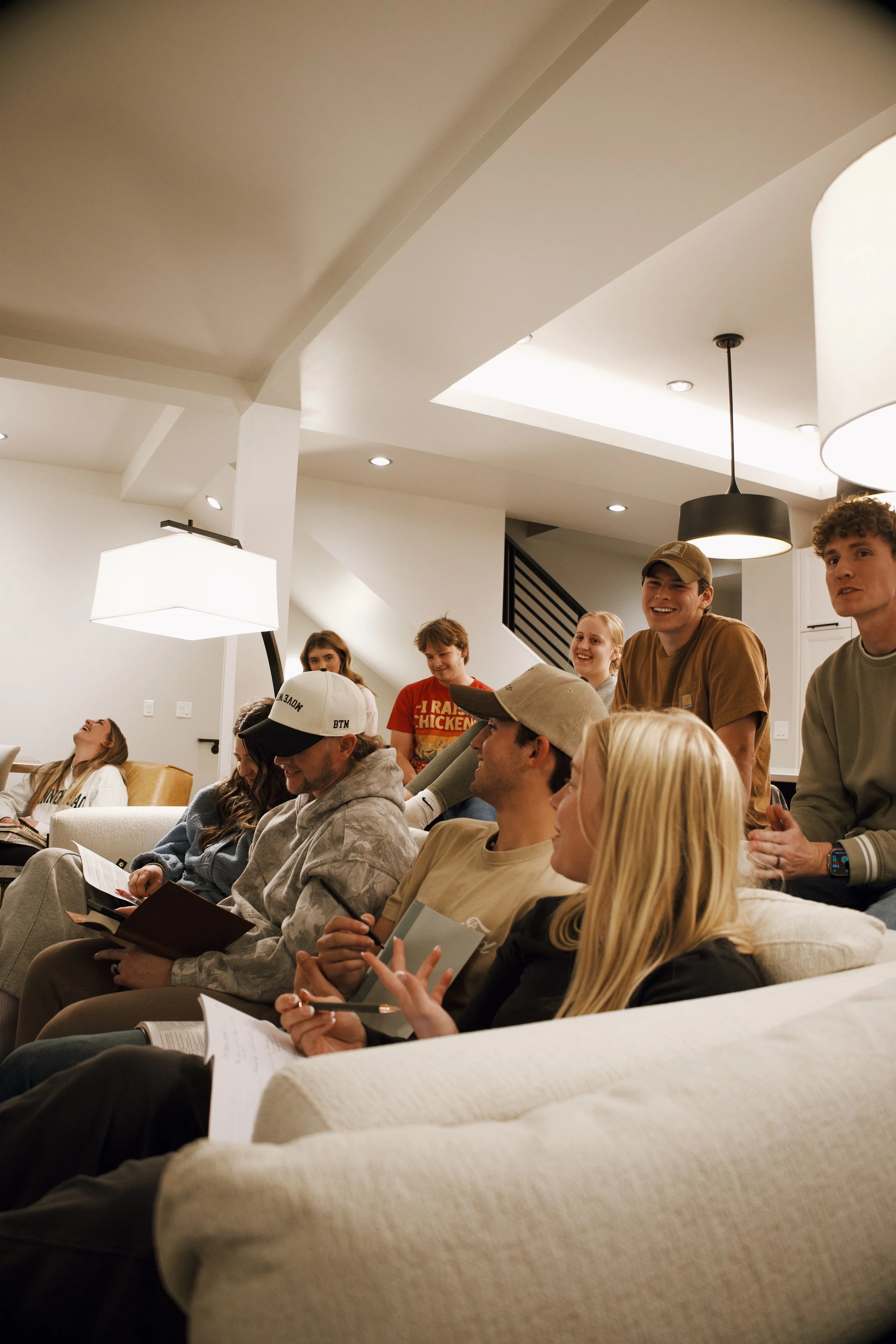 A group of young people gathered in a cozy living room, sitting on couches and chairs, chatting and smiling, with some holding papers and pens.