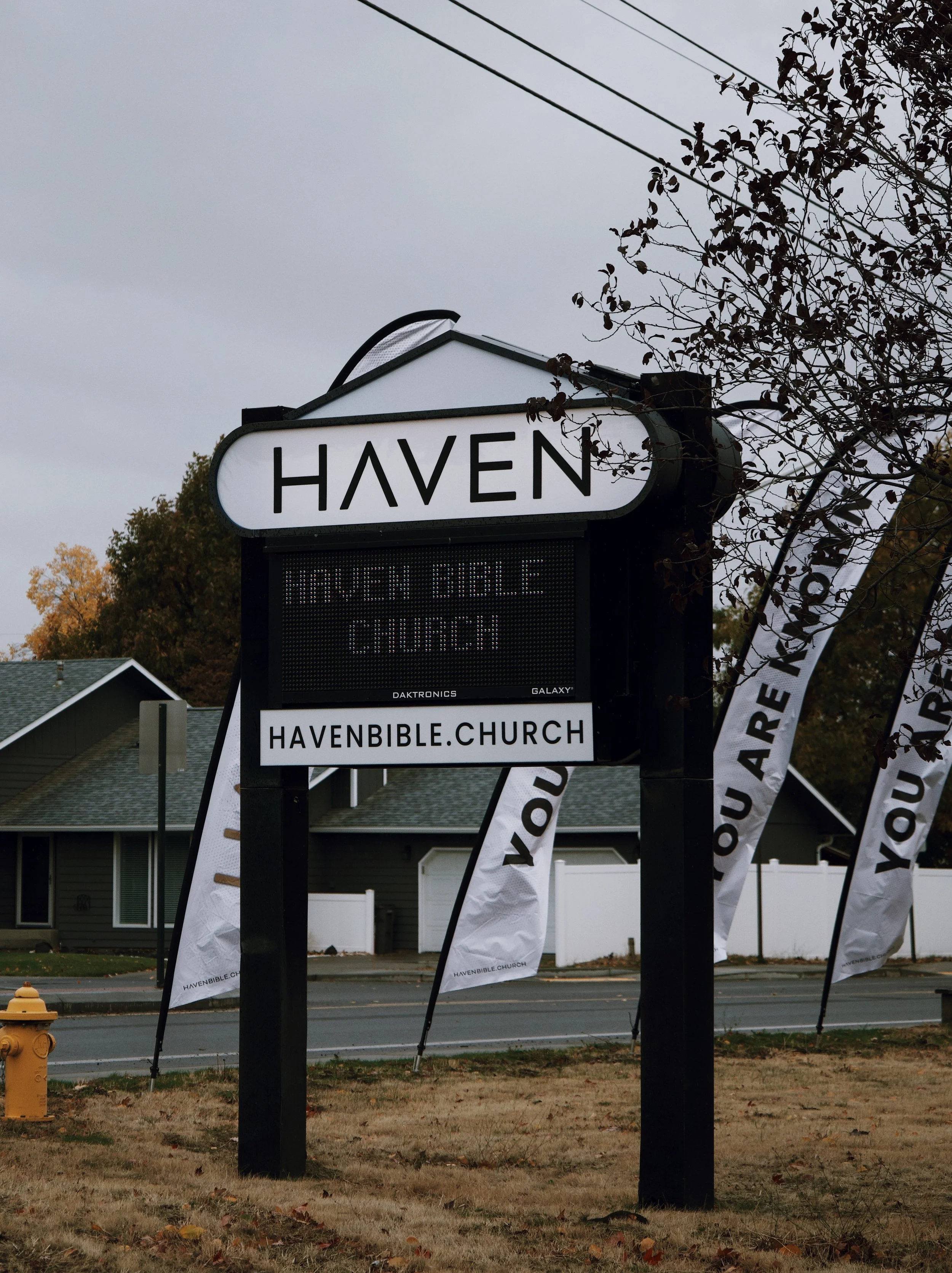 A church sign for Haven Bible Church with the website havenbible.church. Flags with the church's website and message are in the background near a residential neighborhood.