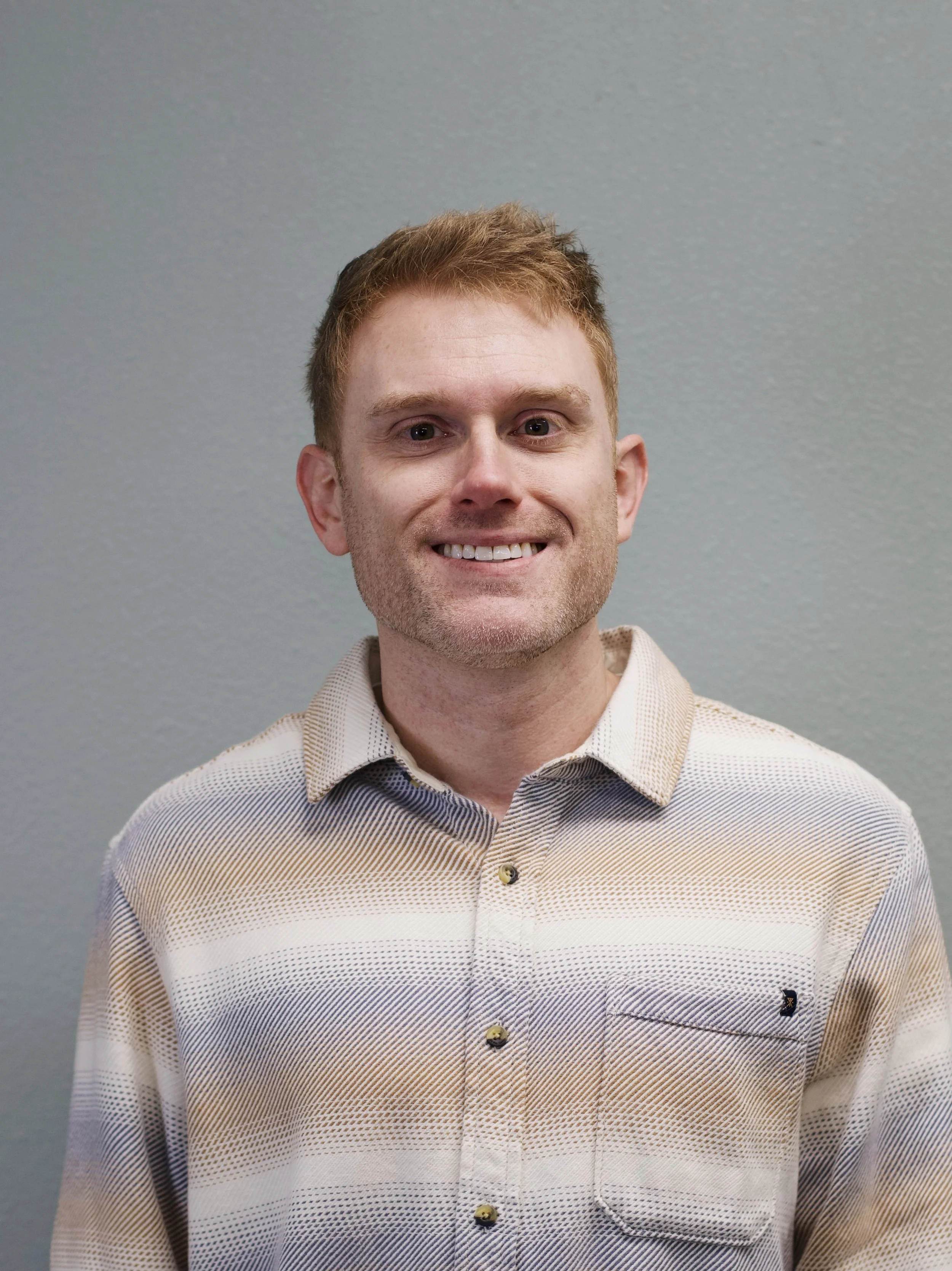 Portrait of a smiling young man with short red hair wearing a gray button-up shirt, standing in front of a red brick wall.