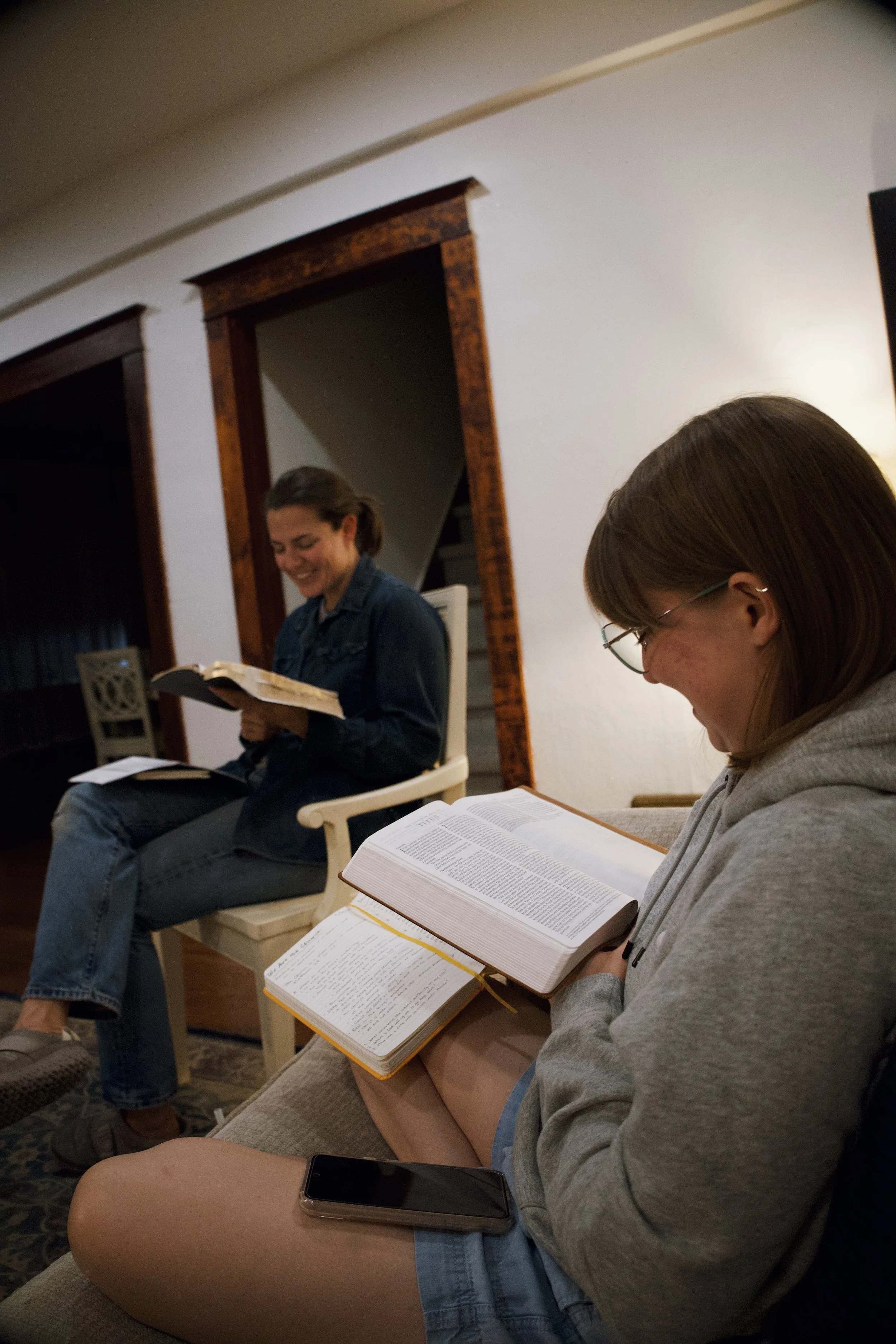Two women sitting on a couch, reading the Bible and smiling, in a cozy living room.