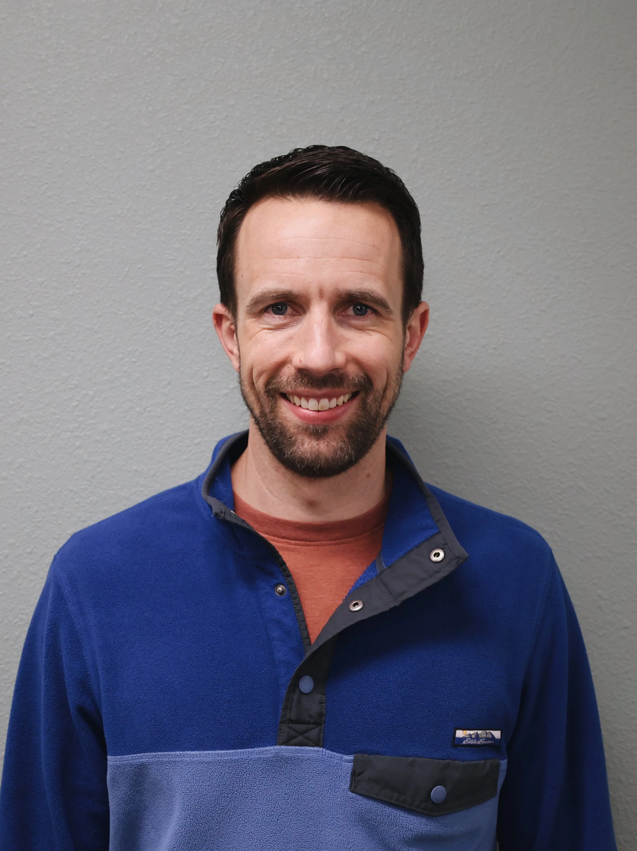 A smiling man with short dark hair wearing a purple button-up shirt, standing against a brick wall.