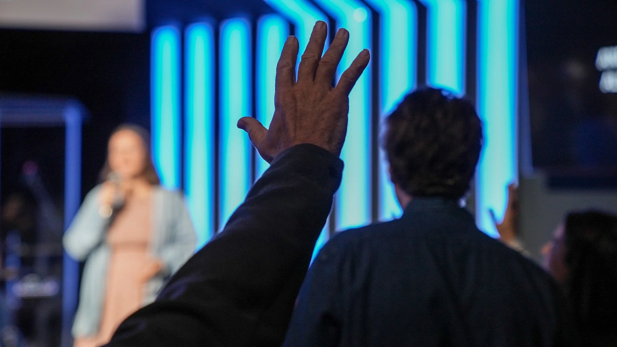 A person raising their hand during a presentation or speaking event with a woman holding a microphone in the background.