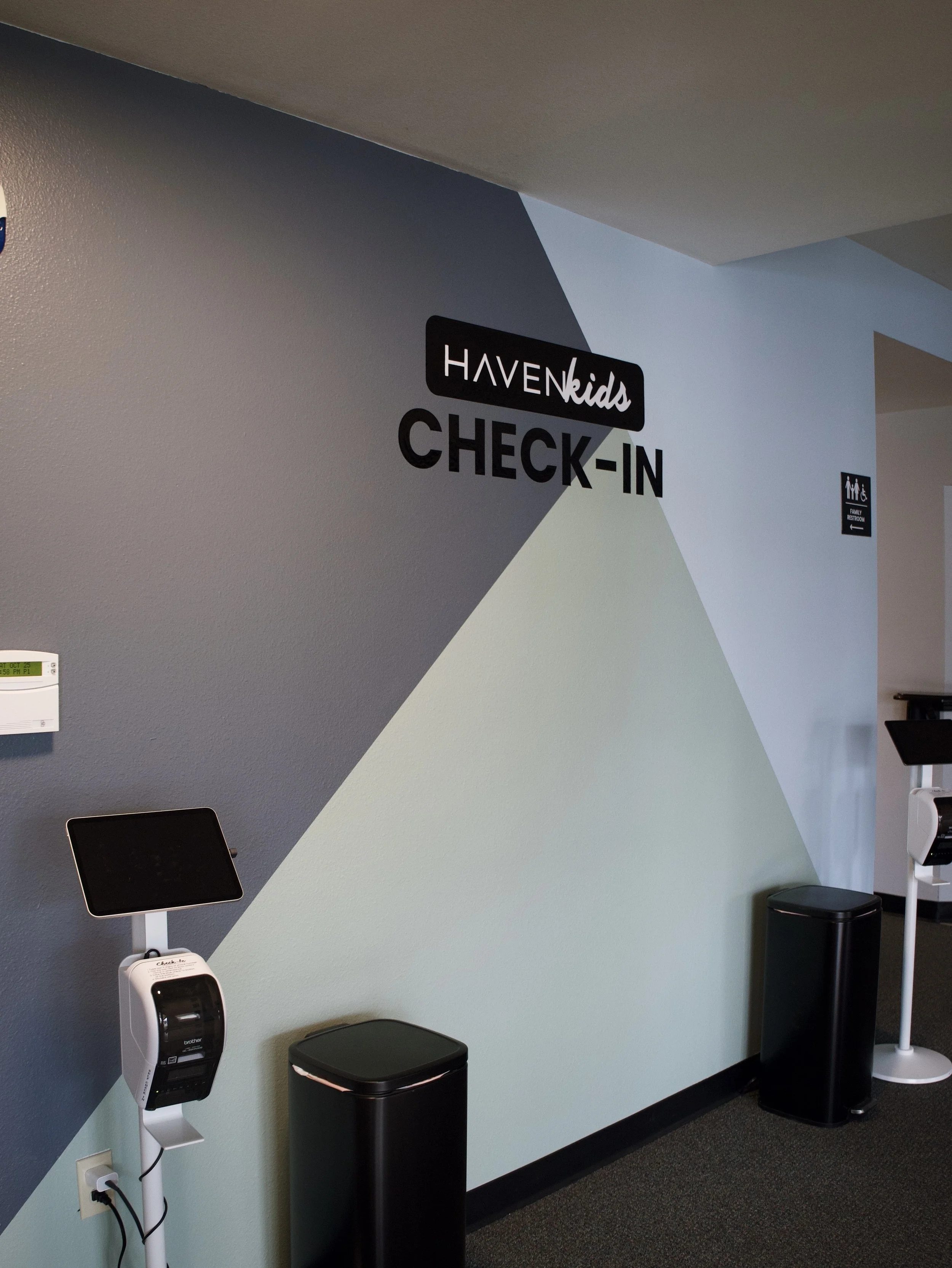 Interior of a Kids' Check-In area with a geometric painted wall, a sign reading "HAVEN Kids CHECK-IN," and two electronic check-in kiosks with touchscreens and printers.