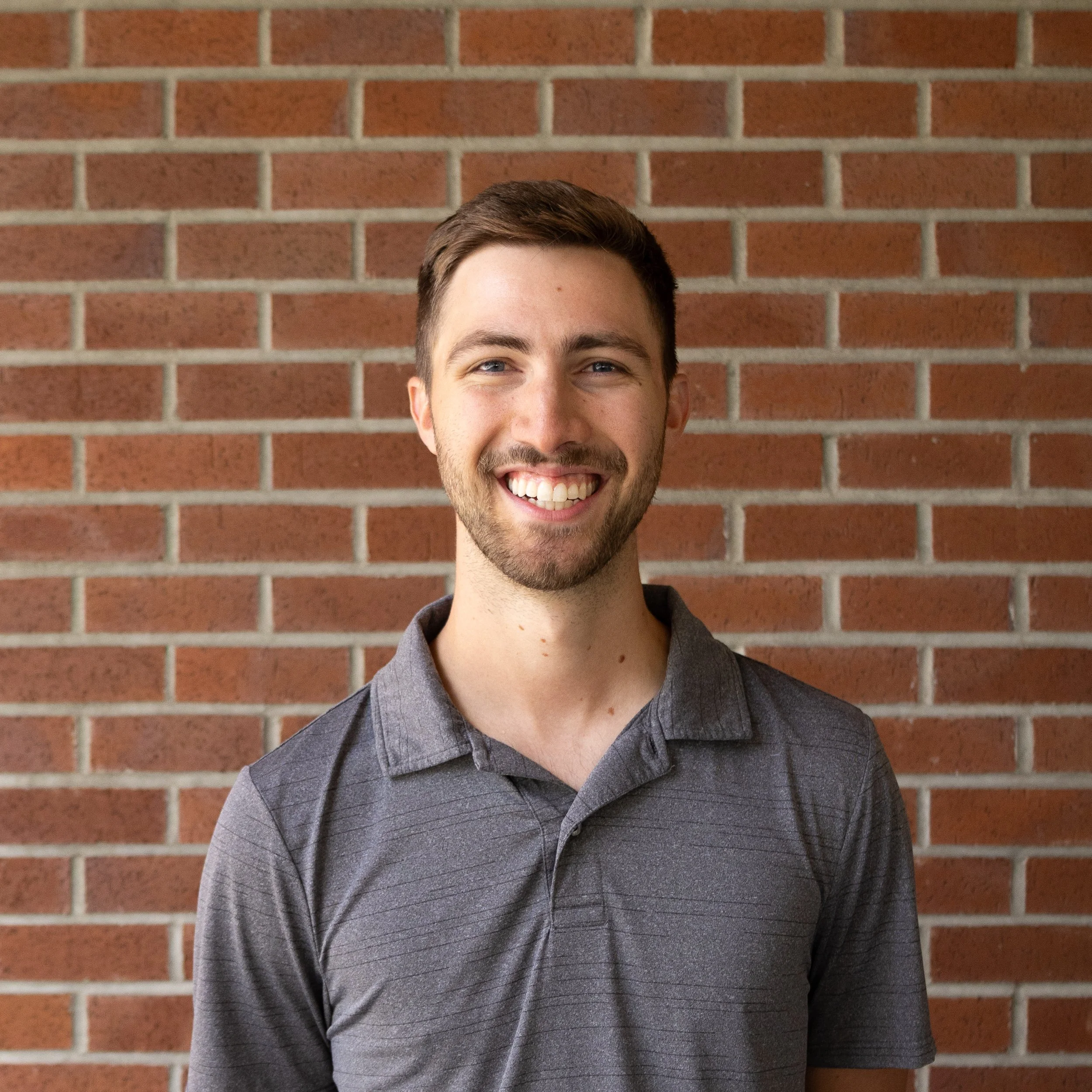 A young man with short brown hair and a beard, smiling, wearing a dark gray polo shirt, standing in front of a brick wall.
