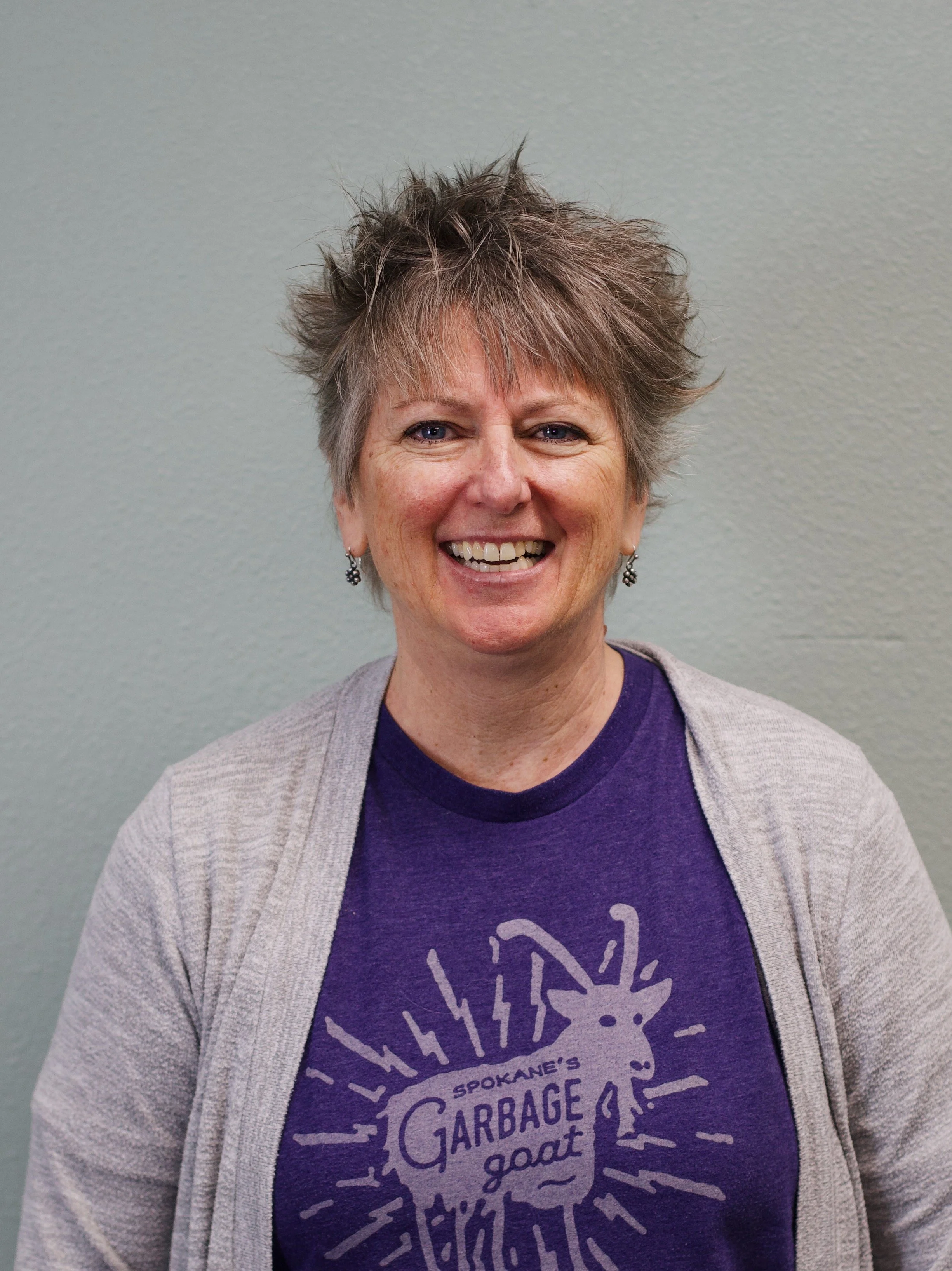 A smiling middle-aged woman with short, spiky gray hair, wearing earrings, a maroon T-shirt with yellow text and a graphic, and a crochet cardigan, standing in front of a brick wall.