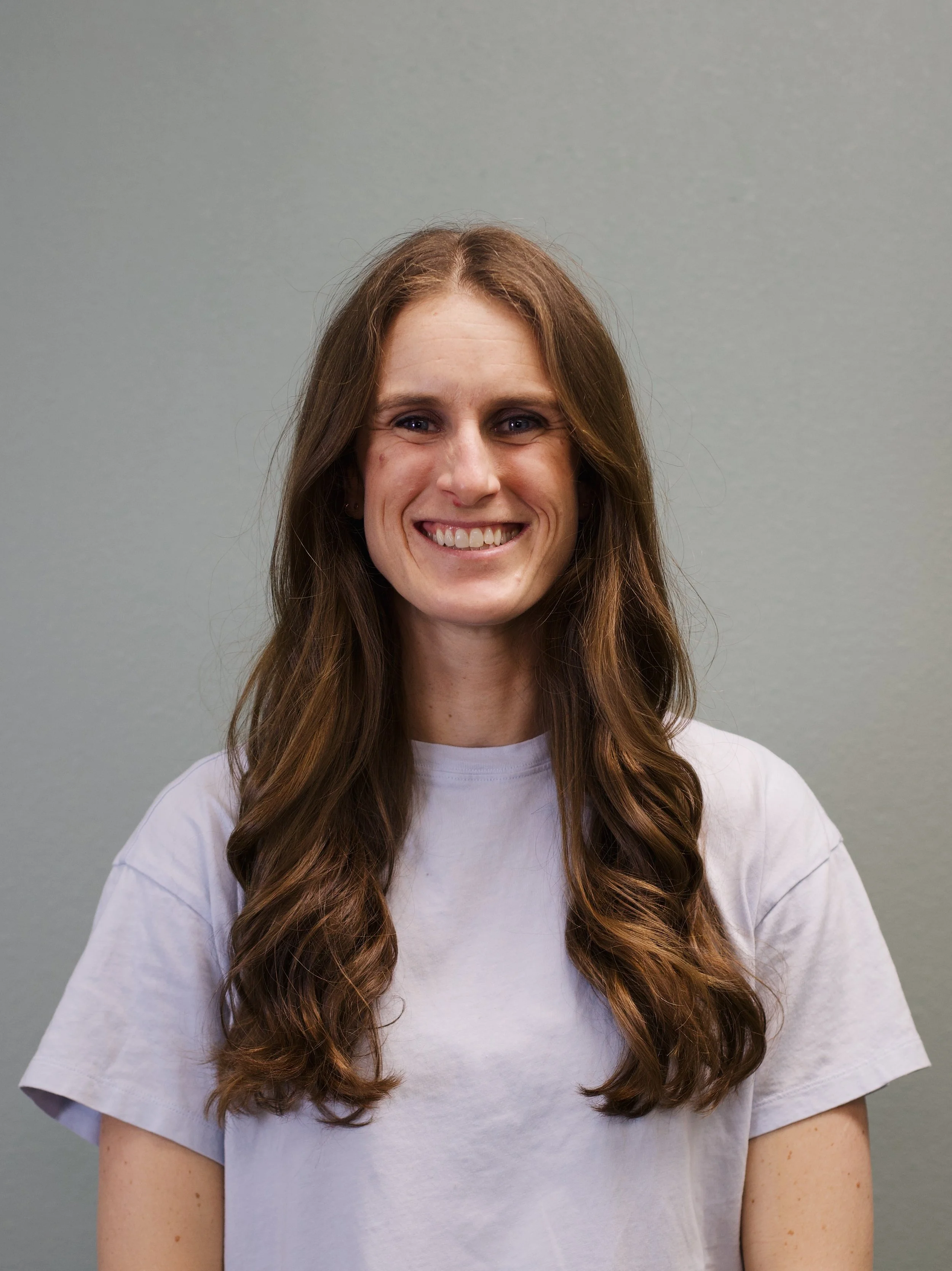 A woman with long, wavy brown hair and a bright smile, standing in front of a brick wall, wearing a black shirt and a gold necklace.