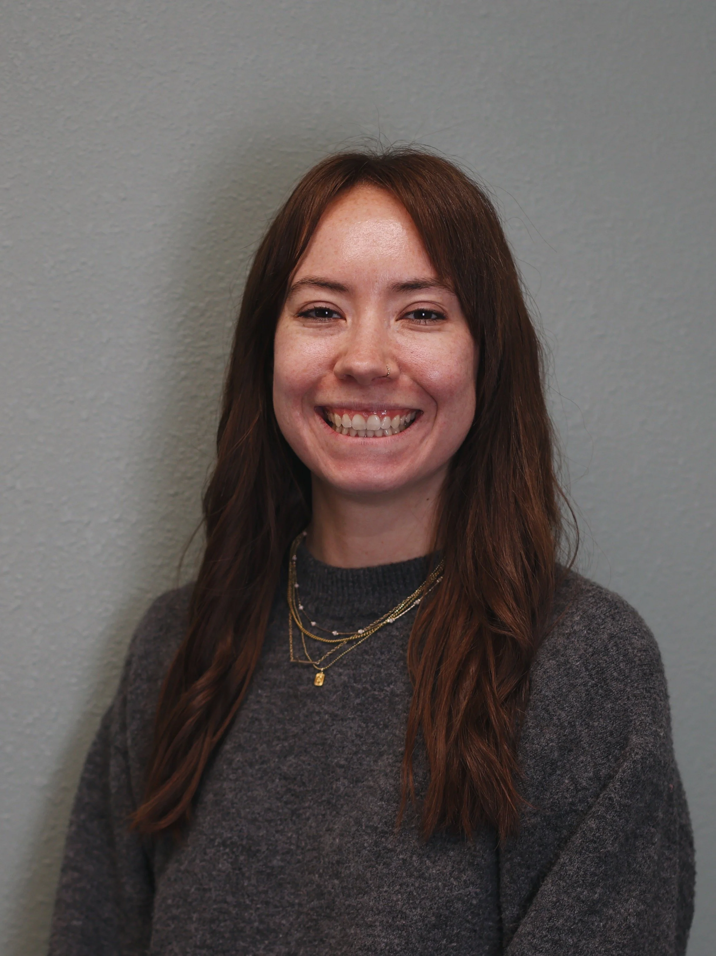 A young woman with wavy red hair and a nose ring, smiling in front of a brick wall, wearing a dark purple shirt and layered gold necklaces.