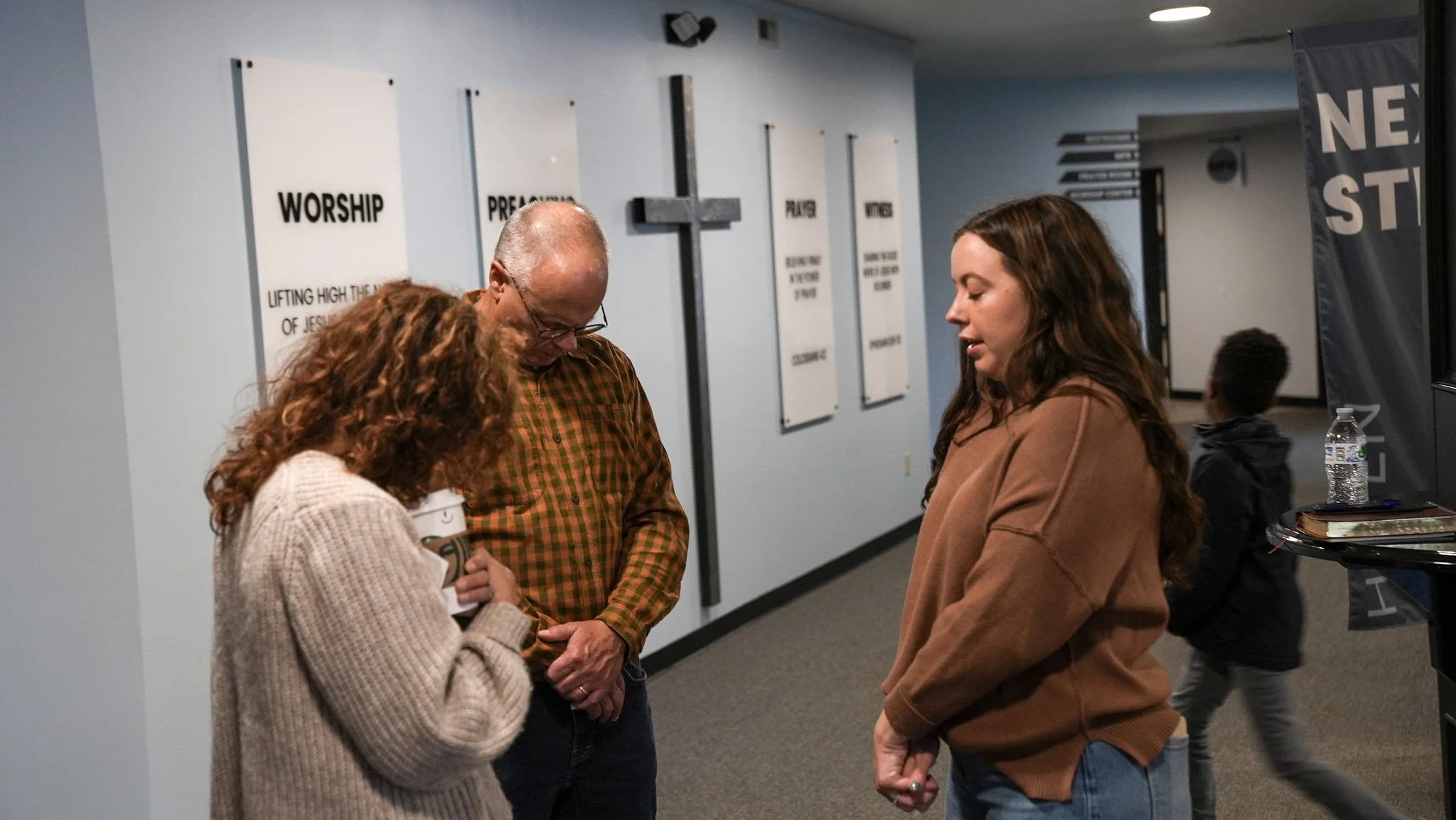 Three people standing together in a church hallway, engaging in prayer. Two women and one man, with children walking by in the background. Church signs and a cross are visible on the light blue wall.